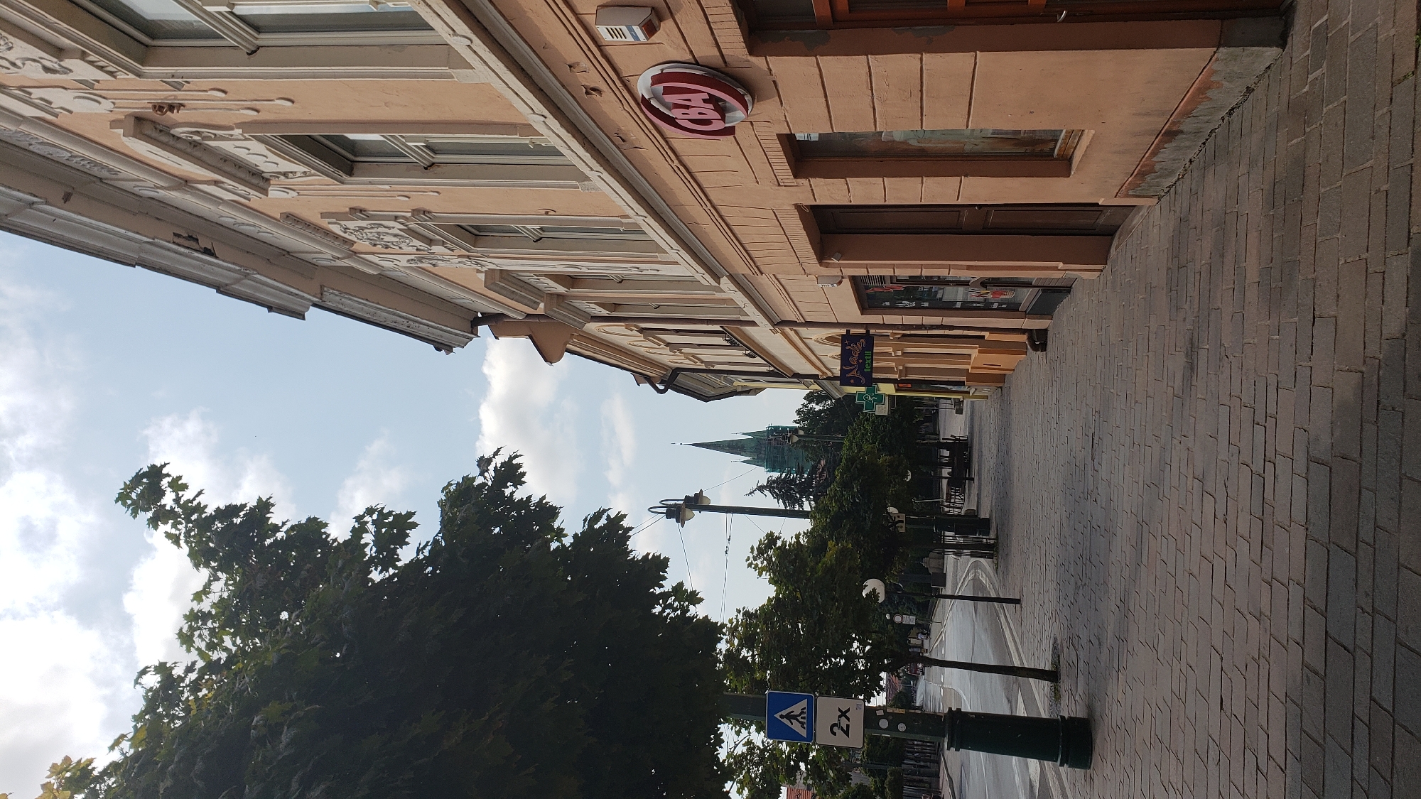The image shows a street scene in the city of Prešov, Slovakia. On the right is the facade of a building with peach-colored walls, ornate architectural details, and windows. A circular red and white sign with the letters "CBA" is visible on the building. Shops are situated at street level with awnings and display windows. A paved sidewalk runs along the building, made of rectangular stone pavers. On the left side of the street, a dark green lamppost stands next to a pedestrian crossing sign with a depiction of a person walking and the number "2x" below it. Mature trees with dark green foliage line the sidewalk and the street. In the distance, a tall, green-spired church steeple is visible against a sky with scattered white clouds. The street appears to be paved, and a few trees are planted along its edge. The overall impression is of a quiet urban street, possibly during daylight hours.