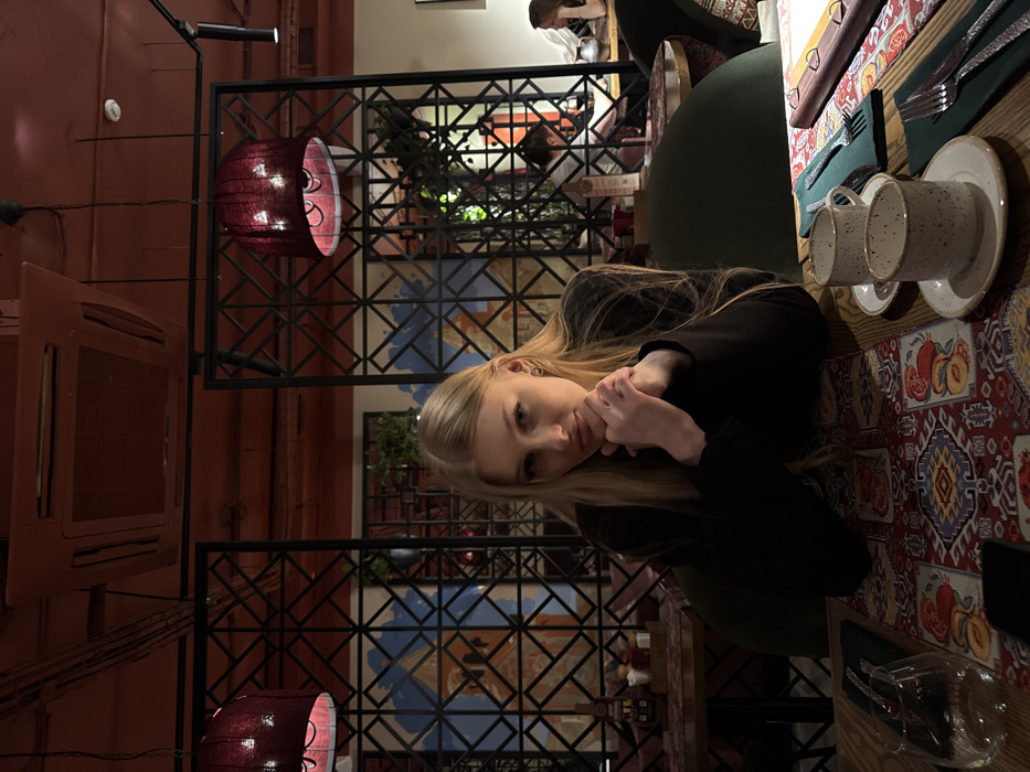 The image depicts the interior of a restaurant or cafe in Perm, Russia. A young Caucasian female with long, straight blonde hair and light skin is seated at a wooden table in the foreground, slightly left of center, facing the viewer. She is wearing a dark-colored, long-sleeved garment. Her forearms rest on the table, with her hands clasped below her chin. A small earring is visible on her left ear.
The wooden table is covered by a patterned table runner featuring geometric designs and stylized fruits in shades of red, orange, blue, and white. On the table to the subject's right are two empty speckled ceramic cups with saucers, and a dark green napkin folded with a fork and knife. A clear wine glass and a dark-colored smartphone are visible to her left. She is seated in a dark green upholstered chair with a curved backrest.
The background shows additional dining areas with other patrons, out of focus. To the subject's right, a person with dark hair is partially visible at a table. Further right, another individual is seen near a large ceramic jug. The restaurant's interior features a dark red ceiling. Two prominent bell-shaped pendant lamps with dark red, ornate shades hang from the ceiling, one on the left and one on the right, providing warm illumination. A square, recessed ceiling-mounted air conditioning unit is also visible. Black metal lattice screens with geometric patterns function as room dividers. Behind these screens, wall art with abstract blue and orange designs, possibly depicting architectural elements, is visible, interspersed with green decorative foliage.