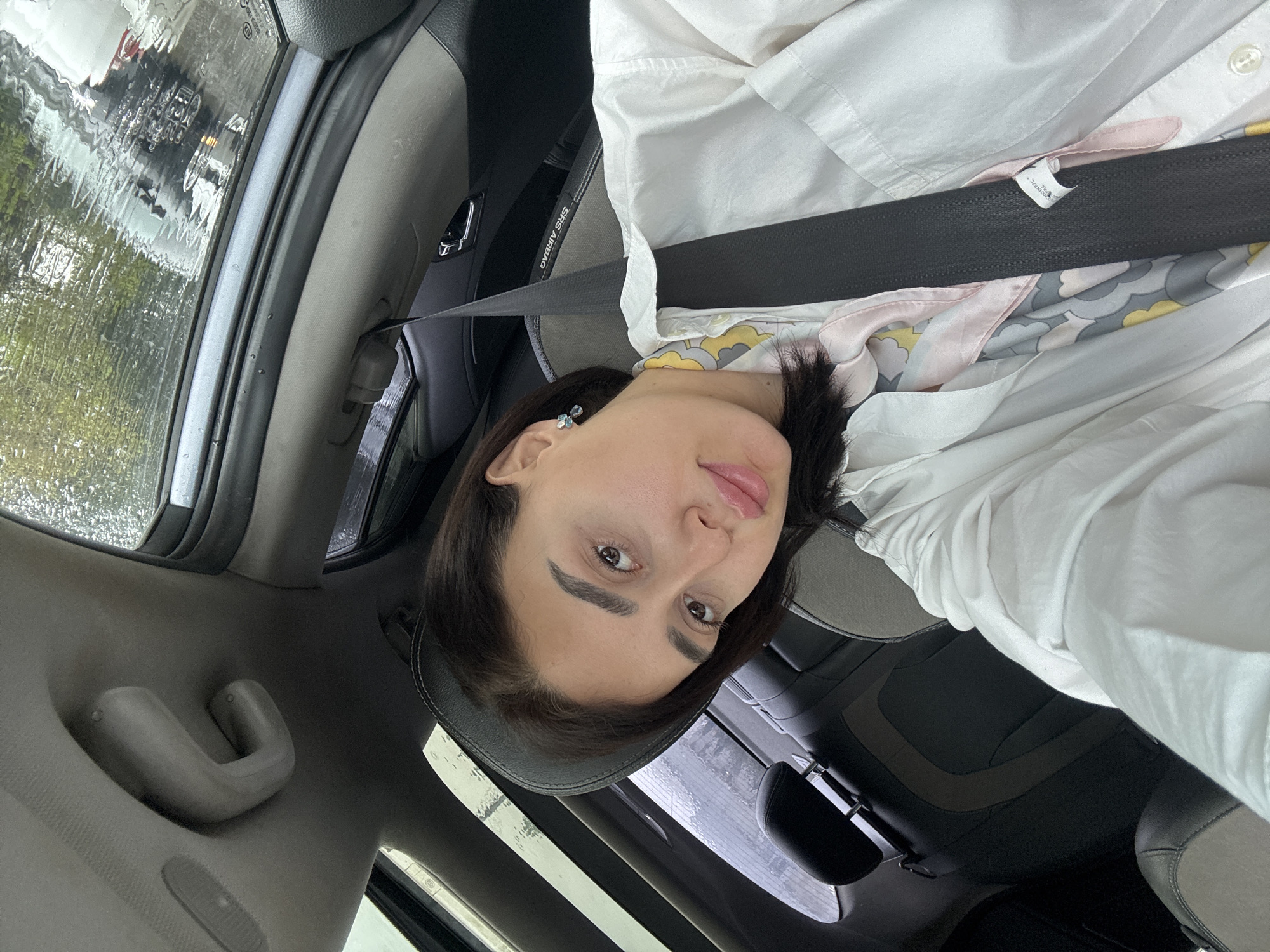 A woman is in the driver's seat of a car, wearing a white collared shirt and a patterned scarf, secured with a black seatbelt. It appears to be raining heavily outside, obscuring the view through the car window. The location is Tbilisi, Georgia. The car's interior is predominantly gray and black. The woman looks directly at the camera.