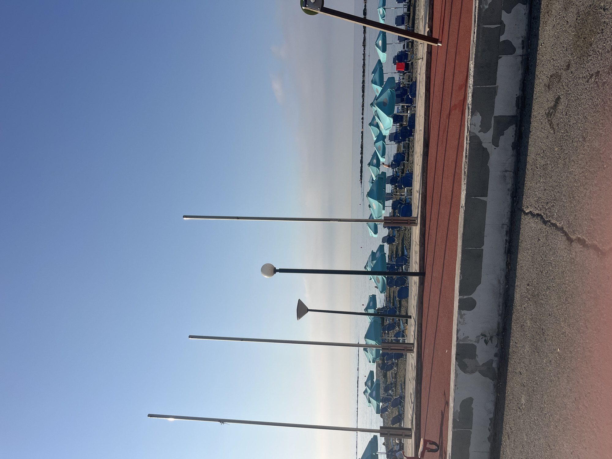 A view of a beach in Agios Tychona Community, Cyprus, on a clear day. In the foreground, a cracked asphalt surface and a red-surfaced pathway are visible, separated by a weathered concrete barrier. Beyond this, a beach with rows of blue sun loungers and turquoise umbrellas stretches towards the calm sea. Several tall, slender poles stand vertically along the beachfront, some with lamps. The sea is a calm blue, meeting a pale sky with a thin layer of haze near the horizon.