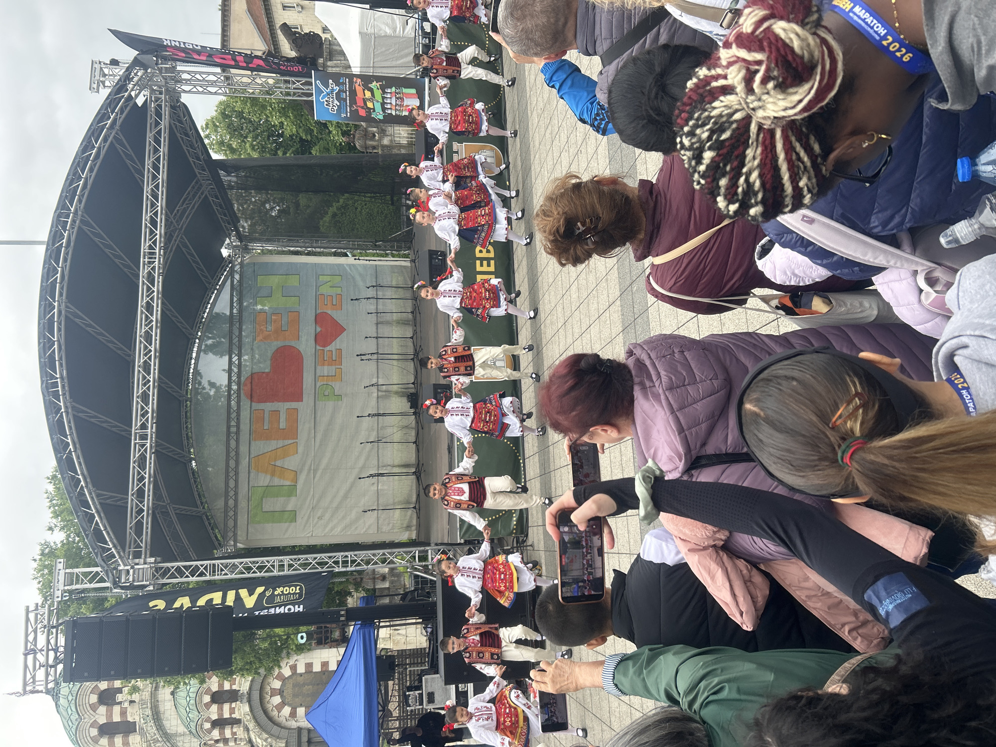 The image captures a scene at a public event in Pleven, Bulgaria, where a group of dancers dressed in traditional folk costumes perform on a stage. The stage has a backdrop with the word "ПЛЕВЕН" (Pleven) prominently displayed. A crowd of people, some of whom are taking photos with their phones, watches the performance. The architecture of the city is visible in the background, including what appears to be a church with a distinctive dome. Banners advertising "VIVA 100% NATURAL HONEST DRINK" are present on the stage. A woman with a lanyard around her neck with the words "МАРАТОН 2026" is visible in the crowd.