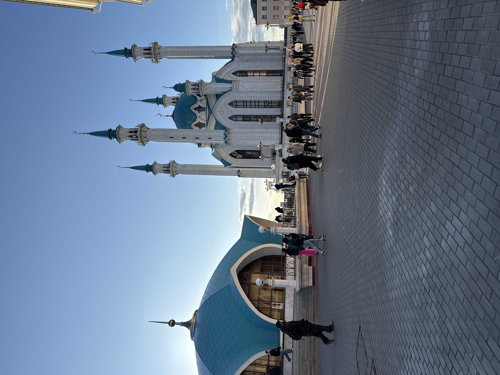 The image shows the Kul Sharif Mosque in the city of Kazan, Russia. The mosque has white walls and blue domes. There are several minarets extending into the blue sky. People are walking and sitting on the brick-paved area in front of the mosque.