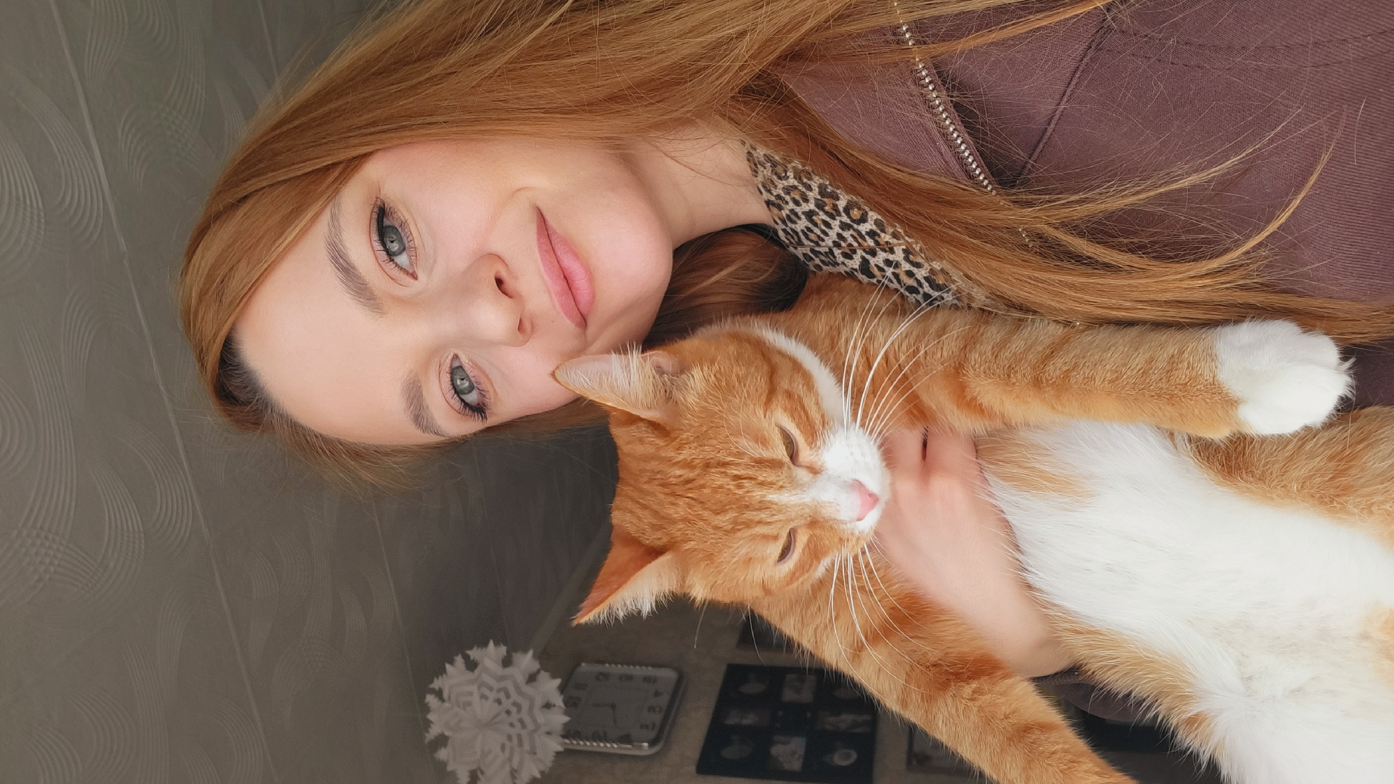 A woman with long, reddish-blonde hair is holding a ginger and white cat. The woman is wearing a brown jacket over a leopard print top. The woman's face is close to the camera, and she is smiling slightly. The cat is resting in her arms, with its eyes closed. The background is blurred, but appears to be an interior setting. A white decorative object is visible in the lower left corner, and what seems to be a clock and a framed photo are also visible in the lower center of the image. The location is Chaikovsky, Russia.