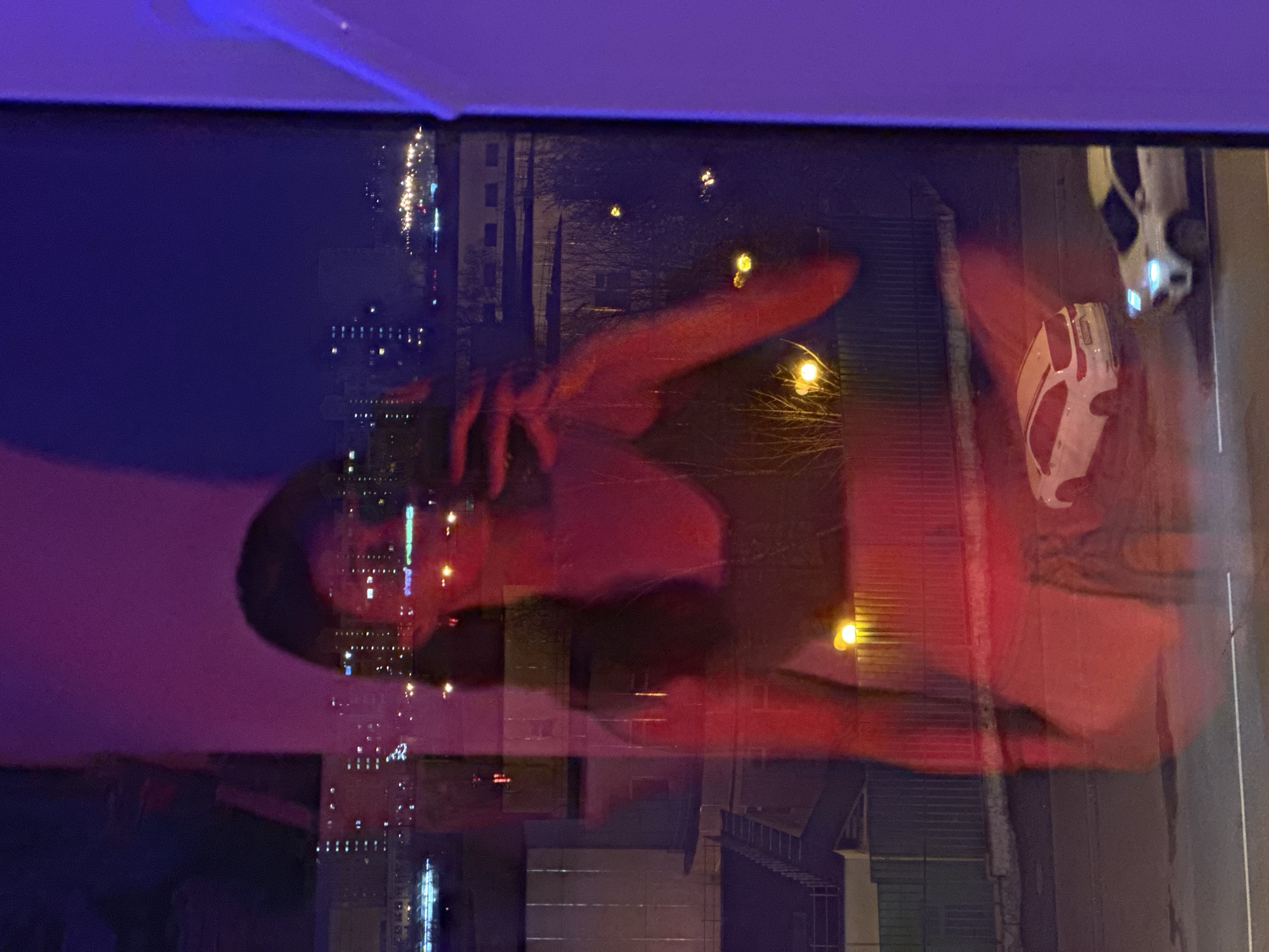 The image appears to show a woman sitting near a window at night. The window reflects the city lights of Moscow, Russia, as well as images of buildings and streetlights. A car is visible outside. The woman's reflection is superimposed on the cityscape, creating a layered effect.