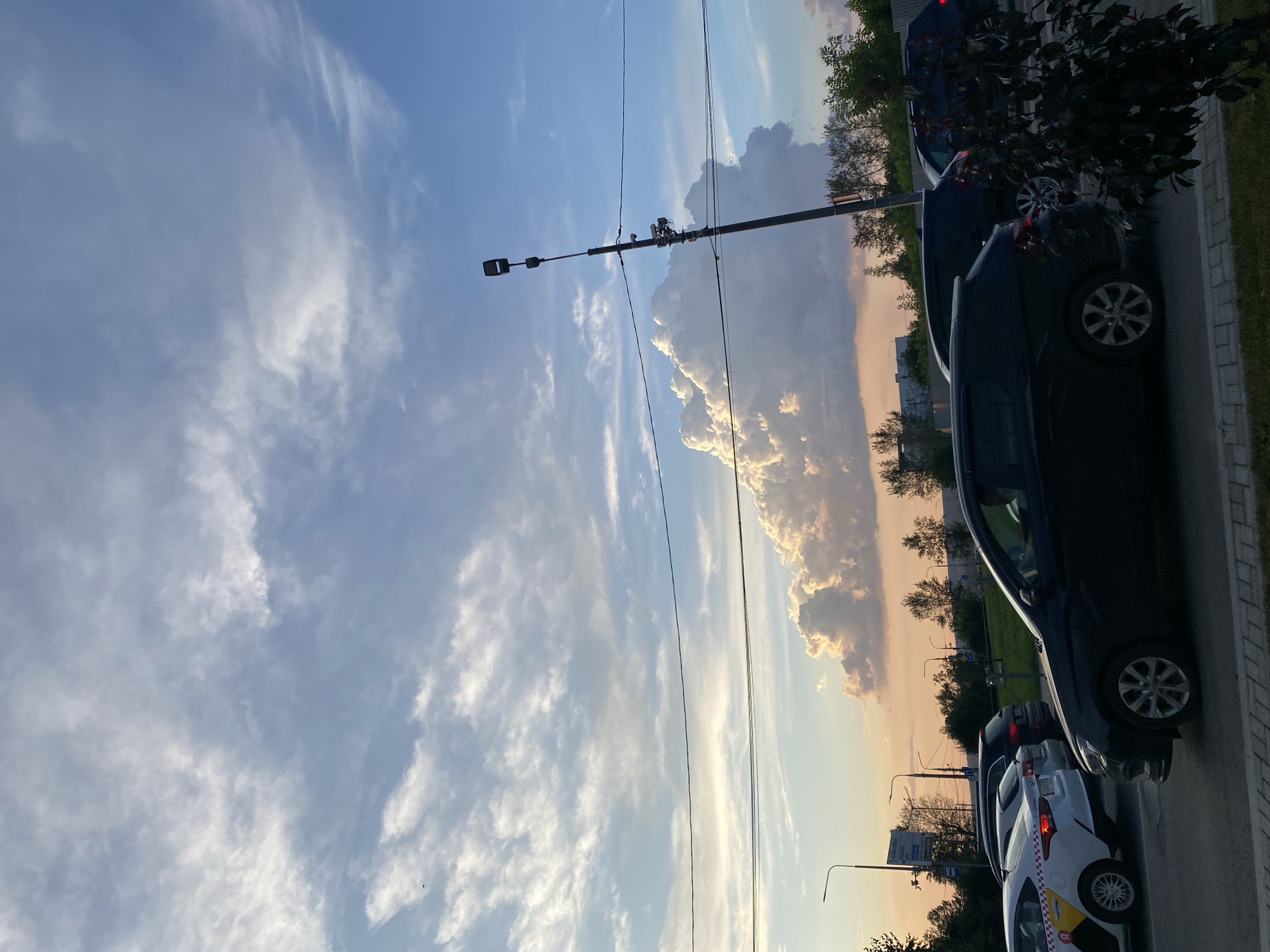 The image captures a street scene with a line of cars in traffic under a dramatic sky filled with clouds. In the foreground, a black car is prominent, with a white taxi visible to its left. The sky is a mix of blue with wispy white clouds and a large, voluminous cloud formation tinged with yellow and orange hues, suggesting it is either sunset or sunrise. A tall lamppost with multiple attachments stands on the right side of the frame, and power lines are strung across the sky. Trees and urban infrastructure, including traffic lights and signs, are visible in the background. The overall impression is a typical urban environment during a period of low light. This scene is located on Lenina Street in the city of Perm, Russia.