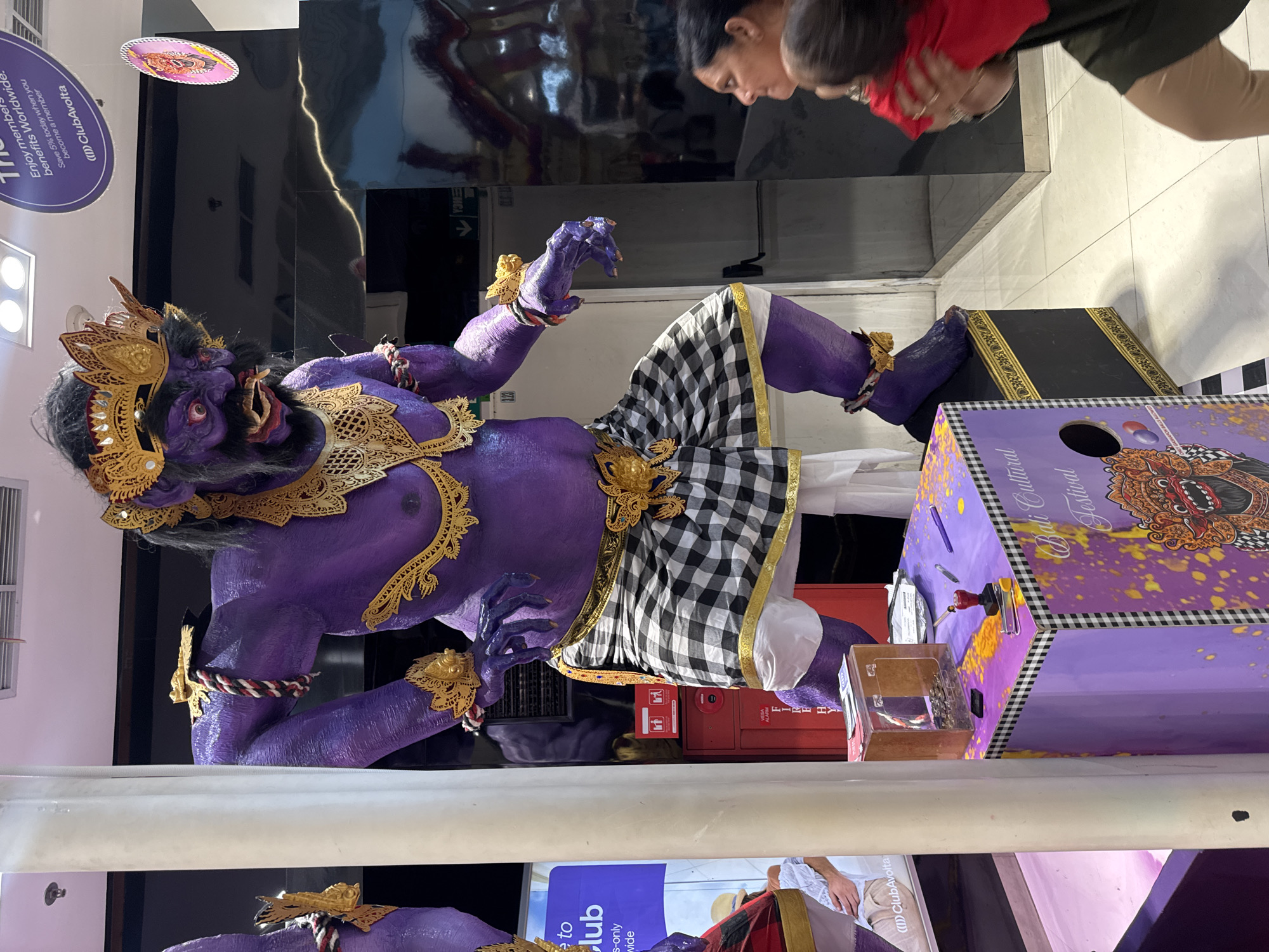 The image shows a large, purple, ornate statue of a demon-like figure adorned with gold accents and a checkered cloth around its waist. The statue is positioned next to a purple box labeled "Bali Cultural Festival", possibly part of an exhibit or display. Two people, an adult and a child, are seen looking at the statue. The location appears to be indoors, possibly in a mall or airport, based on the glossy floors, signage, and other people visible in the background. The mention of "ClubAvolta" suggests the location could be in an airport duty-free area. The statue is the main subject of the image, and the overall scene is likely captured in Kuta, Indonesia, based on the reference to Balinese culture.