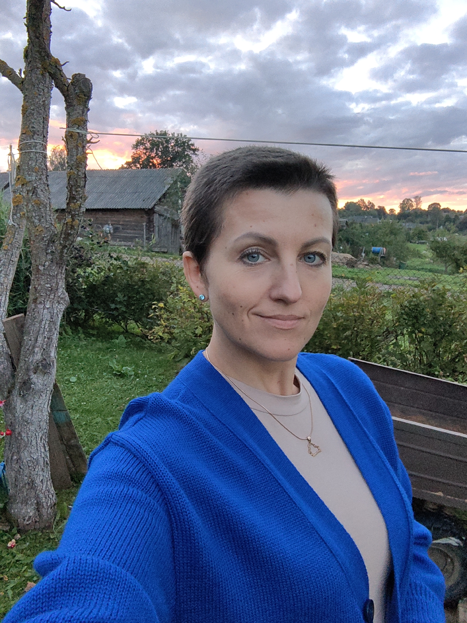 A middle-aged woman with short brown hair and blue eyes is taking a selfie outdoors at dusk. She is wearing a light beige top and a bright blue cardigan. A delicate gold necklace with a heart-shaped pendant rests on her chest. She has a small, light blue stud earring in her left ear. The background shows a rural scene with a wooden building with a corrugated roof, green grass, and overgrown bushes. Two trees stand to the left of the frame, one with visible moss. The sky is partly cloudy, with hints of pink and orange from the setting sun. The overall impression is that of a casual outdoor setting in a village or countryside. The location is identified as Дзямідавіцкі сельскі Савет, Belarus.