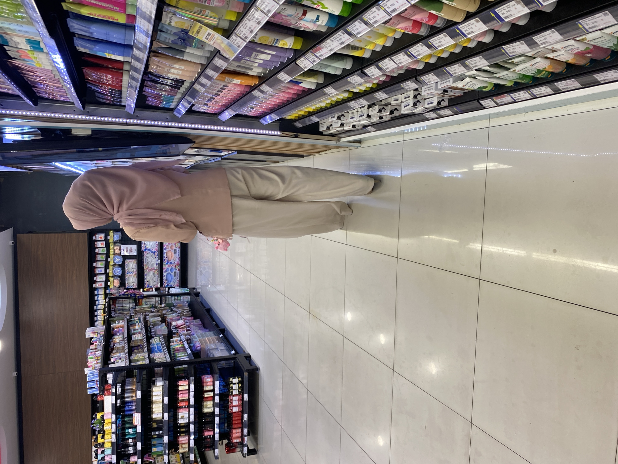 A person wearing a light pink hijab and a light pink jacket over light beige pants is standing in a brightly lit supermarket aisle. The person is facing away from the camera and appears to be looking at a shelf filled with various beauty products, mostly tubes of creams and lotions, arranged in rows. The shelves are black metal and are well-stocked. To the left of the person, there are more shelves containing bottles and boxes of products. The floor is made of large, light-colored square tiles, which are clean and reflect the overhead lighting. The overall environment suggests a modern retail store, likely a pharmacy or a department store section selling personal care items. The scene is located in a supermarket in Banda Aceh, Indonesia.
