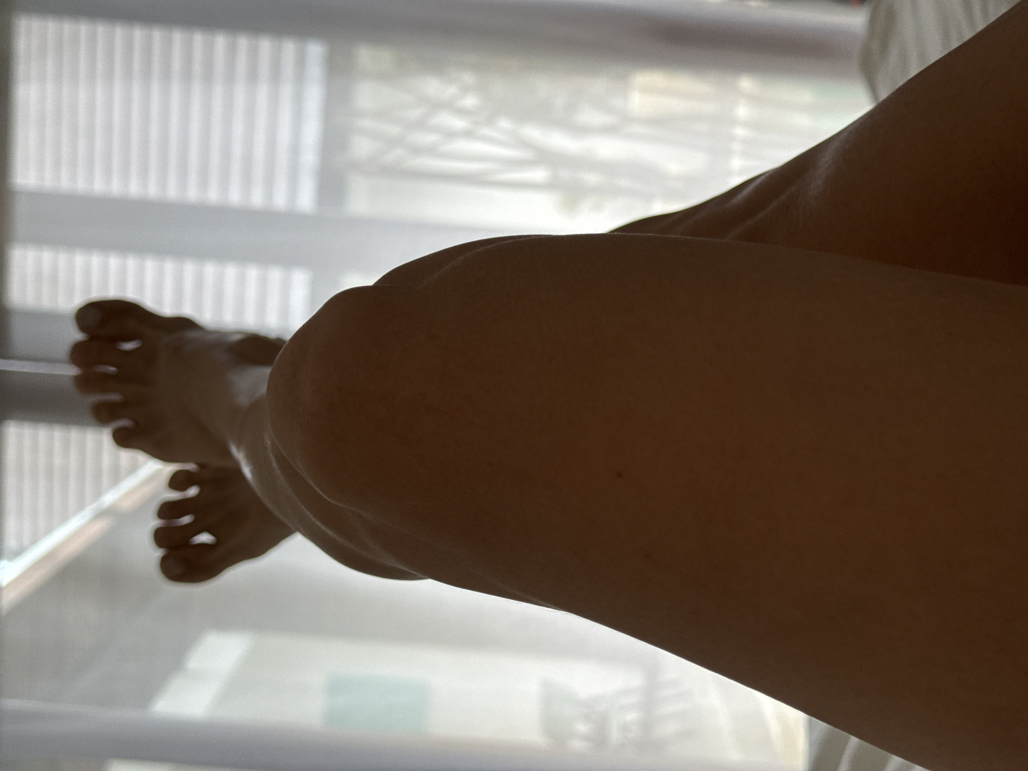 This image presents a close-up view of a person's legs and feet in a relaxed position. The limbs, including the thighs, knees, and calves, are seen from a low angle, with a warm, possibly tanned, skin tone. Soft, diffused natural light illuminates the subject from behind or above, creating a gentle silhouette that emphasizes the contours of the legs. The person's feet are visible on the left side of the frame, with toes subtly curled.
The background shows a window covered by vertical blinds or slats, which filter the ambient light, casting a subtle pattern of light and shadow. The overall lighting suggests it is daytime, likely morning or late afternoon, providing a serene and tranquil atmosphere. The setting is indoors, implying a private space such as a bedroom or living area, where the person is resting. The scene conveys a strong sense of calm and repose. No specific activity is depicted beyond relaxation. There is no visible text in the image. The location context of North Kuta, Indonesia, is consistent with the relaxed, warm environment suggested by the image.