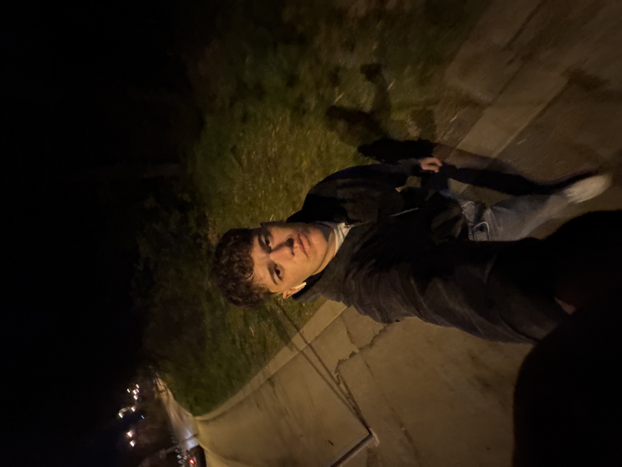 A medium shot shows a young man in jeans and a dark jacket on a sidewalk at night. He appears to be wearing an earpiece. A patch of grass is visible behind him, and in the distance, lights suggest a city street. While the details are limited due to the low light, the context suggests an urban environment, possibly Sofia, Bulgaria.