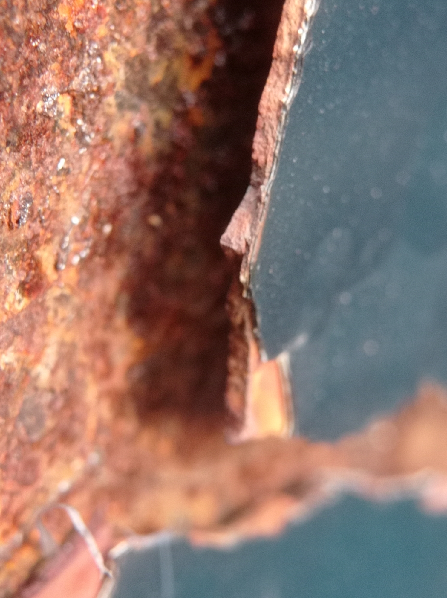 The image shows a close-up, macro view of rusted metal and a blue surface. The rusted metal appears to be a piece of an object, possibly part of a vehicle or structure, with rough, flaky textures in shades of brown and orange. Some reflective specks are visible on the rusted surface. The blue surface, which fills a significant portion of the right side of the image, is smooth and appears to be painted metal or plastic. It has a slightly matte finish with some small, out-of-focus circular highlights, suggesting it might be a car or a piece of equipment. The scene is entirely focused on the textures and materials, with no discernible context of location within Mulyoagung, Indonesia, or any indication of people or actions.