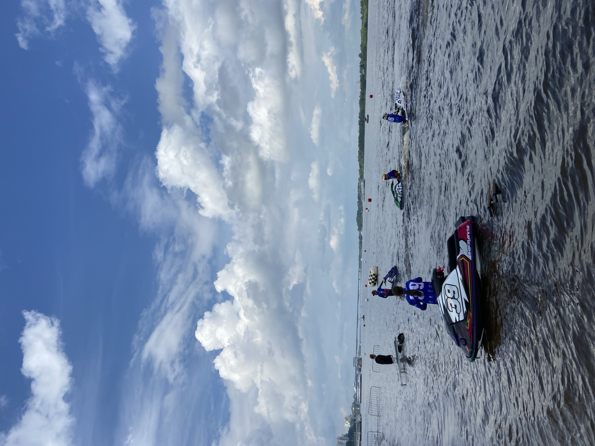 An outdoor, medium shot captures a water sports event on a river under a bright, cloudy sky, possibly in the city of Perm, Russia. The foreground features a jet ski with the number 39 displayed prominently, partially submerged in the rippling water. In the midground, several individuals on jet skis are engaged in the activity, some in motion and others stationary. One person stands near the water's edge with what appears to be a trailer. The background reveals a city skyline with buildings and a bridge spanning the river, suggesting an urban setting. The water is choppy, and buoys mark the course for the racers. The sky is a mix of blue and white clouds, with some larger, dramatic cloud formations.