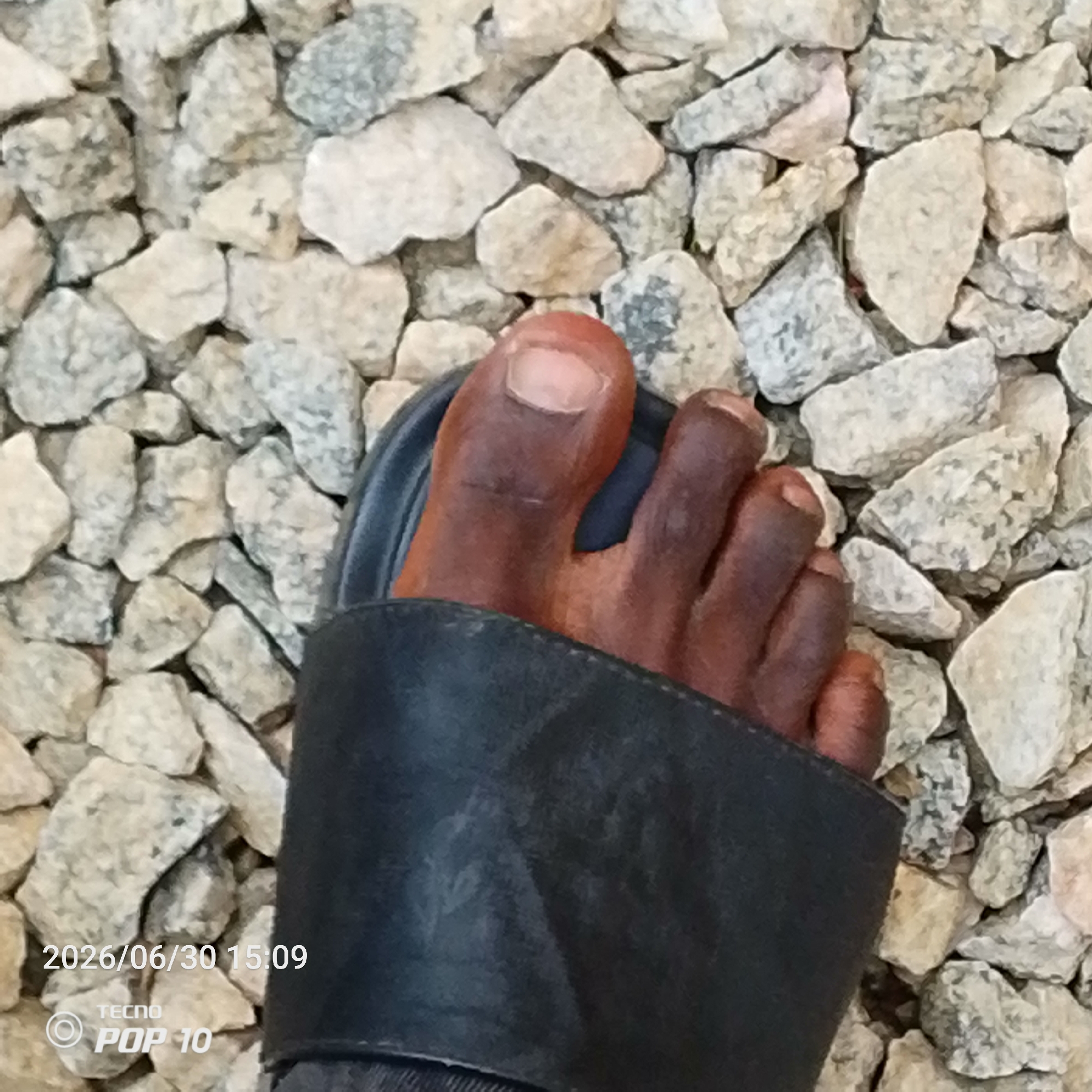 A close-up, overhead shot focuses on a person's dark-skinned foot wearing a black sandal, positioned on a bed of light-colored gravel. The foot is angled slightly to the right, with toes clearly visible and a portion of the sandal strap covering the arch. The gravel appears to be small, irregular stones of varying shades of gray and beige, creating a textured surface. The lighting suggests an outdoor environment, with no discernable shadows indicating the time of day. A timestamp in the bottom left corner reads "2026/06/30 15:09" and a device logo "TECNO POP 10" is also present. This scene is consistent with a location in Jalingo, Nigeria, where such gravel surfaces are common in outdoor areas and public spaces.