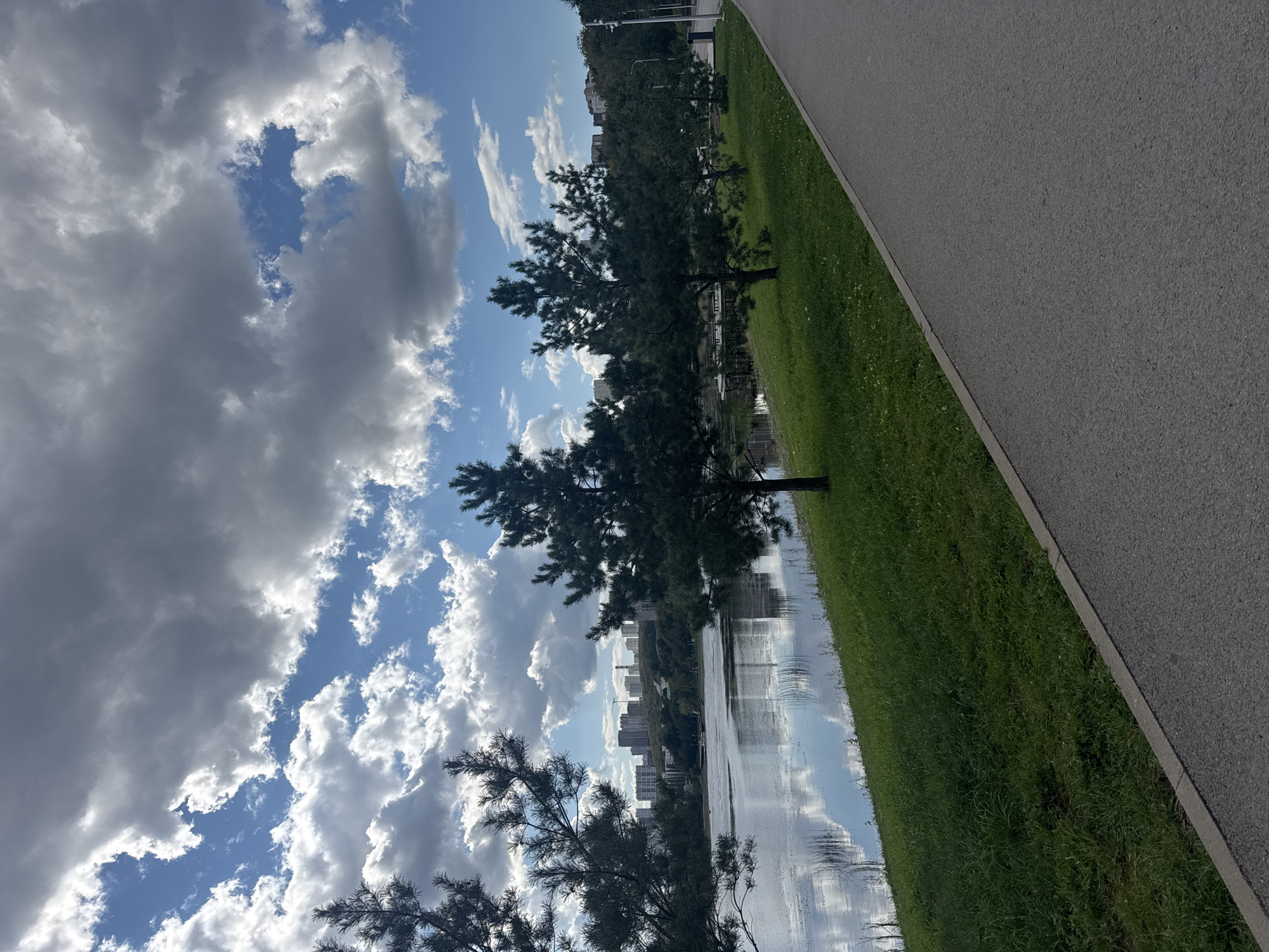 This image depicts a scenic view of a lakeside park with a paved walkway. In the foreground, the walkway curves to the right, bordered by a grassy slope leading down to the water. Several coniferous trees are spaced along the grassy area. Across the calm lake, a line of multi-story buildings, characteristic of urban development, stretches across the horizon, with some cranes visible, indicating ongoing construction. The sky is filled with a dynamic mix of dark storm clouds and bright, sunlit cumulus clouds, suggesting a partly cloudy or changeable weather condition. The lake reflects the sky and the distant cityscape. The location is identified as the park along the Ufa River in Ufa, Russia. No people are visible in the image.