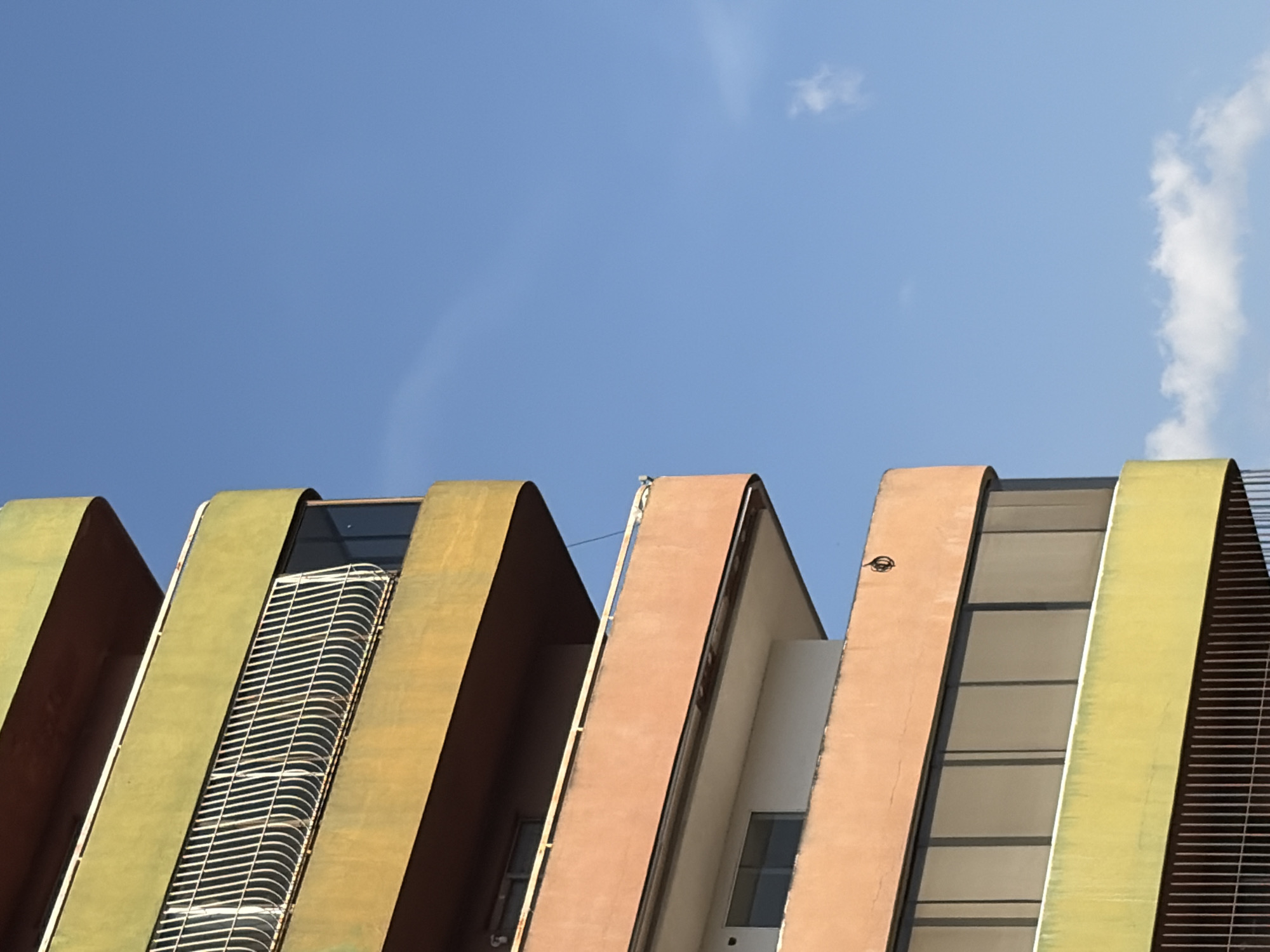 The image presents a striking architectural detail of a building under a clear, bright blue sky with wispy white clouds. The perspective is an upward-looking shot, emphasizing the unique design of the structure. The building consists of several parallel, wave-like sections, each with a distinct curved facade that extends upwards and then curves back inward, creating deep channels between them.
These facades exhibit a vibrant palette of earthy tones, primarily olive green, burnt orange-brown, and a lighter yellowish-green. The surfaces appear textured, possibly weathered or with a stucco finish. Between the curved exterior walls, vertical sections of windows, glass panels, and metallic grates are visible. On the leftmost section, a prominent white slatted grate or screen, which also features a slight curve, covers an opening or window. Further right, some sections reveal darker glass windows and lighter interior walls. A small, dark, circular object, possibly part of the building's infrastructure like a light fixture or small antenna, is visible attached near the top edge of one of the orange-brown facades.
The lighting indicates a sunny day, likely midday or early afternoon, with clear skies and strong, direct light highlighting the curves and textures of the building. No human activity or events are depicted; the scene is a static study of modern architecture. The building is located in Ciudad Guayana, Venezuela. There is no discernible text visible in the image.