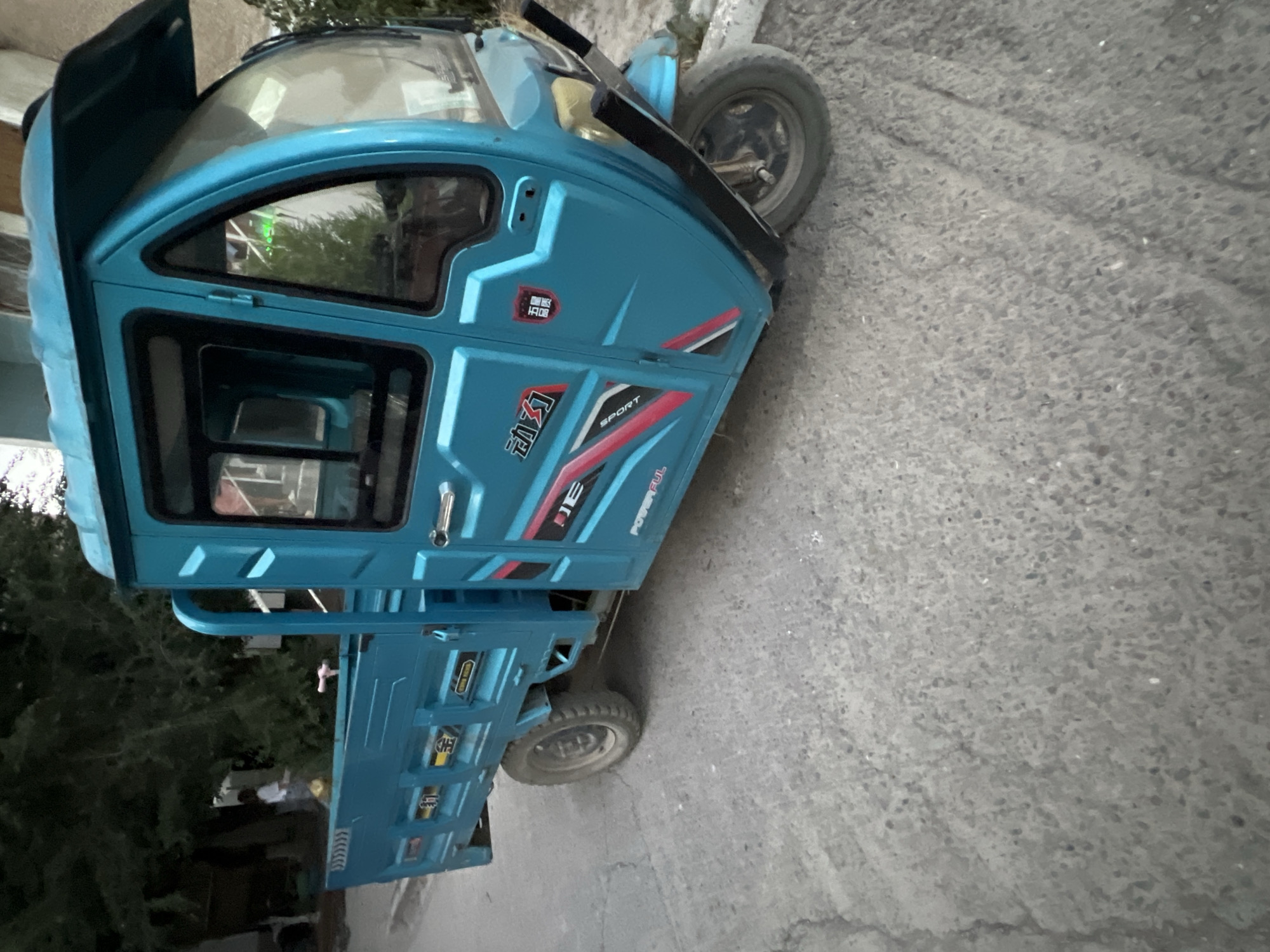 A bright blue, three-wheeled cargo scooter is parked on a paved road. The scooter has a covered cab with windows and a cargo bed in the rear. The side of the cab features a red, white, and black stripe design with Chinese characters and the word "SPORT". The front of the scooter has two headlights and a bumper. A person in a white shirt is visible in the background, standing near some trees. The environment appears to be an outdoor urban setting, possibly in Tokmok City, Kyrgyzstan, given the context.