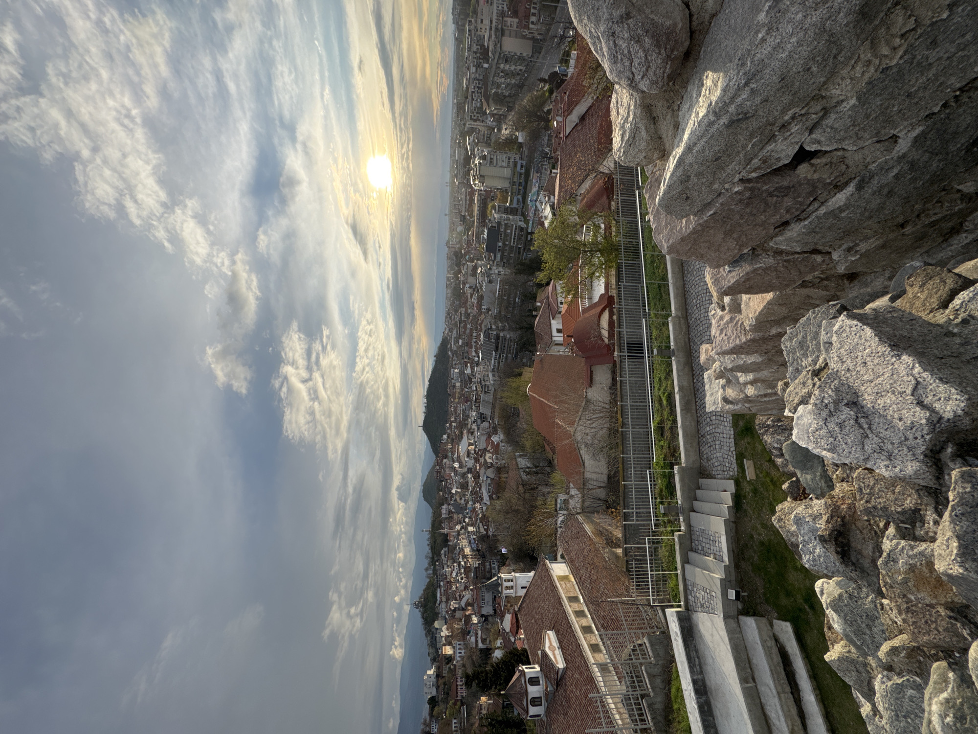 The image shows a view of Plovdiv, Bulgaria, from Nebet Tepe. The perspective is from above, looking down towards the city at sunset. The foreground includes large, jagged rocks, a metal fence, and concrete stairs. The middle ground shows a dense urban area with red-tiled roofs, buildings of various sizes, trees with bare branches, and a few patches of green. In the background, there is a line of distant mountains and a dramatic sky with the sun setting, creating a bright glow amidst the clouds.