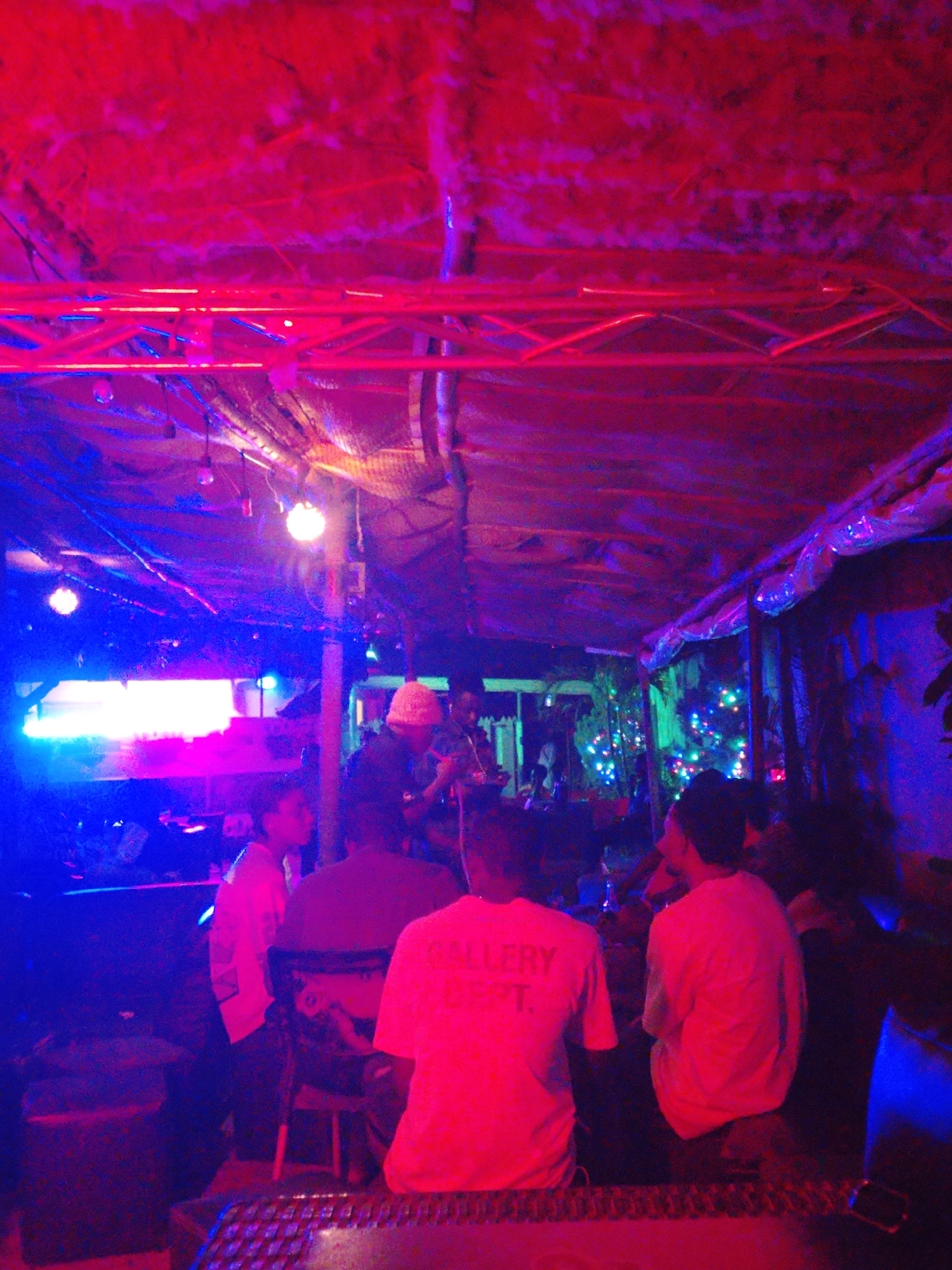 A crowded bar in Unknown, Unknown is illuminated with red and blue lighting, where a live band performs on a stage. Several patrons are seated at tables, watching the performance. One patron is wearing a white shirt with the words "GALLERY DEPT." on the back. A Christmas tree with multicolored lights is visible in the background. The ceiling is low and covered with some kind of material. Above the band, there are metal structures that appear to be part of the lighting system.