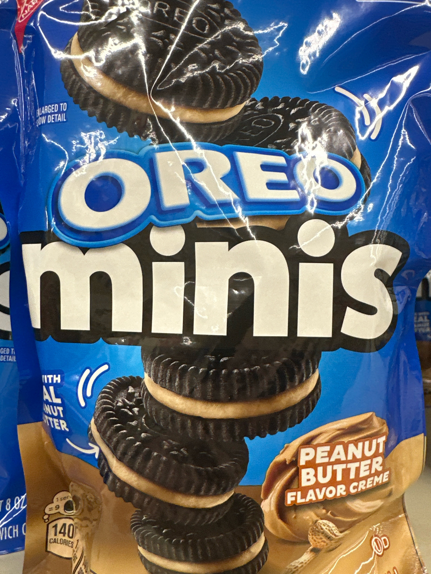 A close-up shot of a bag of Oreo Minis Peanut Butter Flavor Creme cookies. The bag is predominantly blue with the Oreo logo prominently displayed in white and blue lettering. The words "OREO" and "minis" are very large and centered on the bag. Several cookies are stacked on the bag, showcasing their dark chocolate wafers and creamy peanut butter filling. A graphic of peanut butter and a peanut is also visible on the bag. The image appears to be taken in a retail setting, possibly a grocery store aisle in Camp Hill, United States of America, as suggested by the context of product packaging. No people are visible in the image. The lighting is bright and appears to be artificial, highlighting the glossy surface of the plastic packaging.
