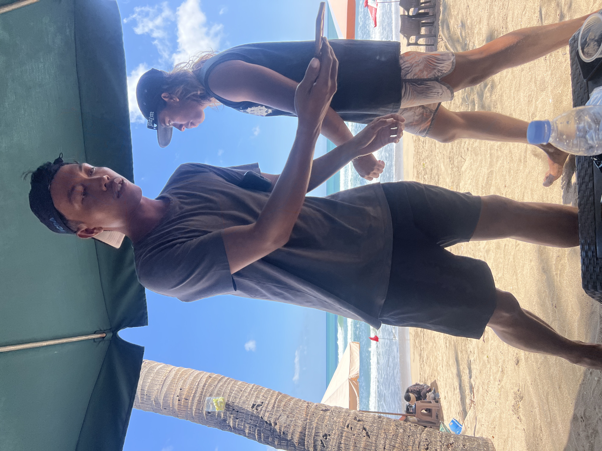 Two young men are standing on a sandy beach in Kuta, Indonesia, under the shade of a large green umbrella. The man in the foreground, wearing a dark gray t-shirt and black shorts, is reaching out to the man behind him. He has a black baseball cap on backwards. The man in the background, wearing a dark sleeveless shirt and patterned shorts, is walking away from the camera and looking to his left. He is also wearing a black baseball cap. Behind them, the turquoise ocean stretches to the horizon under a bright blue sky with scattered white clouds. A palm tree stands to the left of the frame, and beach umbrellas and chairs are visible further down the beach.