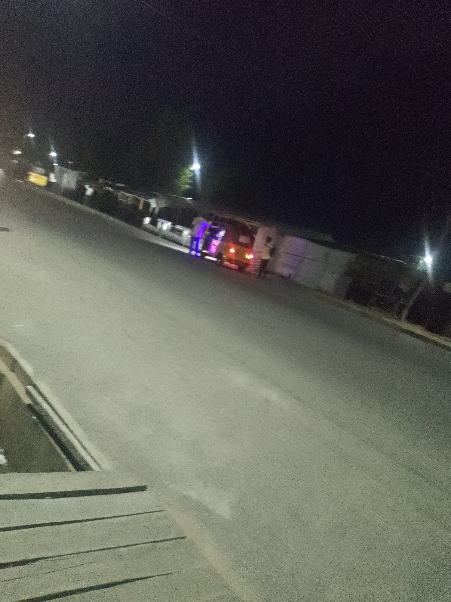 At night, on a paved road in Jalingo, Nigeria, a yellow van is stopped near a white structure. Two individuals are near the van, one wearing a light-colored shirt and dark pants, the other wearing a dark top and light pants. The van has its hazard lights on, indicated by flashing red lights at the rear. The scene is illuminated by streetlights and the vehicle's lights, creating a dark environment with some visible light sources. The road surface is asphalt. The image captures a static moment with no clear indication of movement or interaction beyond the presence of the people and the stopped vehicle.