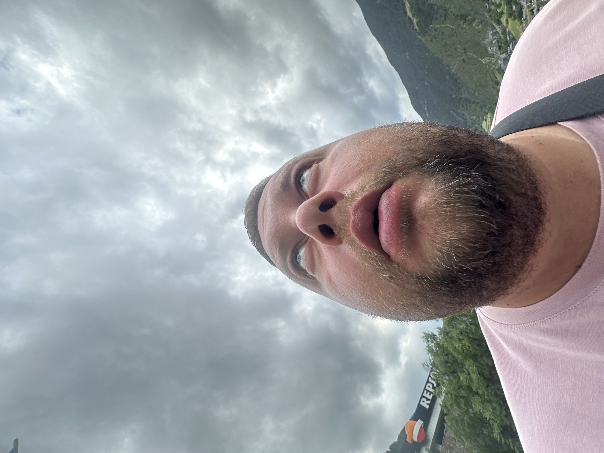 The image is a close-up, rotated view of a man's face and upper torso, captured from a low angle looking upwards. The man has a beard and mustache, and is wearing a light pink t-shirt with a dark shoulder strap. His lips are slightly parted, and his left eye is visible, gazing intently upwards.
The background features a heavily overcast sky filled with numerous grey clouds, indicating dull weather during the daytime. In what would be the upper right if the image were correctly oriented, a dark, rugged mountain covered in green foliage rises, with a small village or settlement faintly visible on its slopes. Towards the bottom right, amidst green trees, a "REPSOL" sign is partially visible on a building or structure, displaying black letters on a white background along with its distinctive red and orange shell logo.
The man's activity appears to be taking a selfie, utilizing an extreme upward perspective that highlights his face against the cloudy sky and the towering mountain landscape. The visible text is "REPSOL". The scene depicts a mountainous environment, consistent with the location context of Encamp, Andorra.
