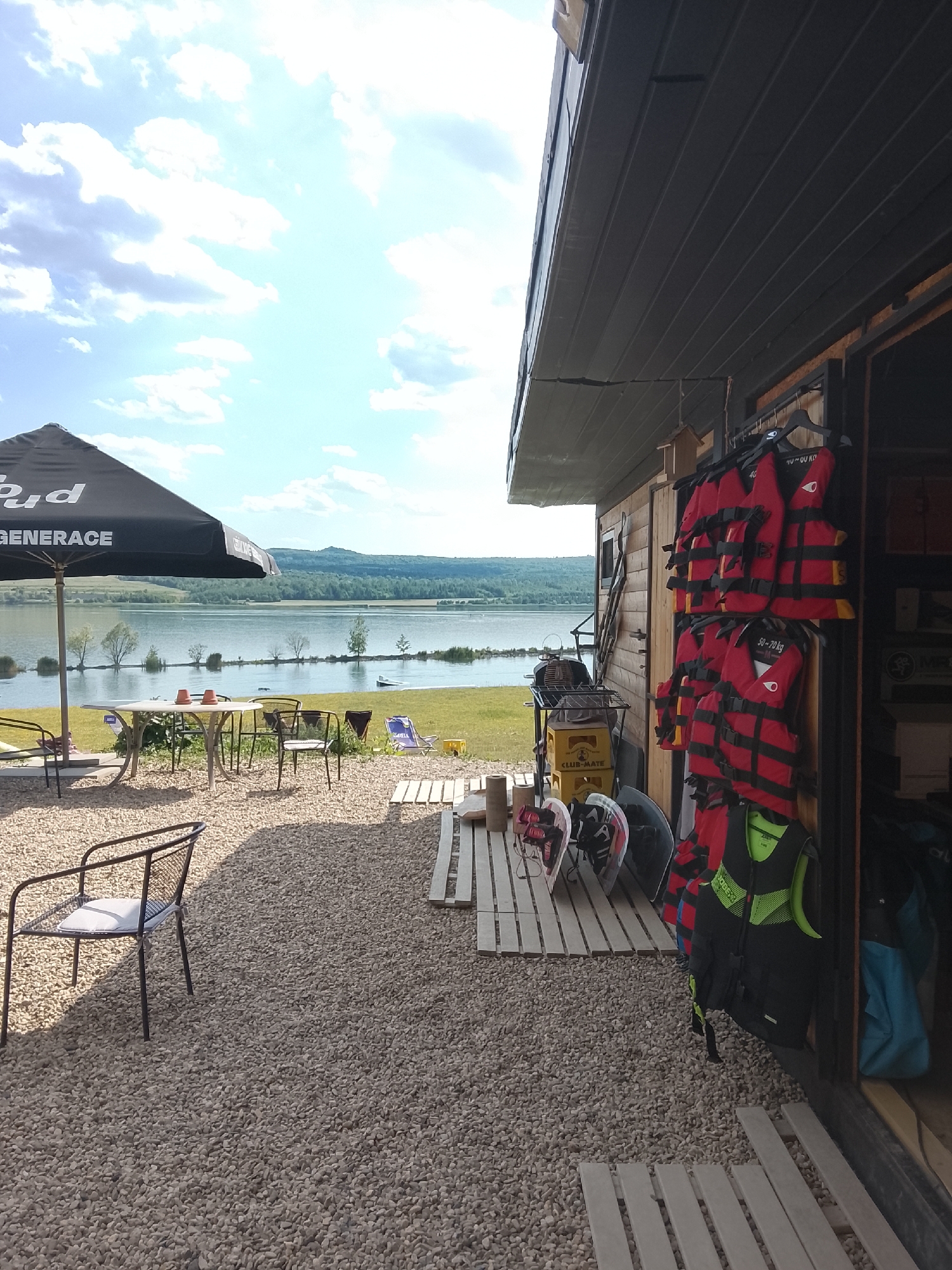 This media file captures an outdoor scene with a focus on water sports equipment and a recreational area by a lake. The main subjects include several red and black life vests hanging on a rack outside a wooden structure, suggesting a rental or service area for activities like kayaking or paddleboarding, as indicated by the presence of paddleboards leaning against the wall. A large black umbrella shades a table with chairs in the foreground, creating a relaxed seating area. Beyond this, a calm lake stretches to the horizon, bordered by trees and rolling hills under a bright, partially cloudy sky.
The scene appears to be set during the daytime, likely late morning or early afternoon, given the strong sunlight casting shadows. The weather seems pleasant, with clear skies and a gentle breeze implied by the open-air setting. The location appears to be a lakeside recreational facility, possibly a club or park, offering access to water activities. There are no people actively engaged in activities in this specific frame, but the setup suggests preparations for or a lull in such events.
Notable details include the text "GENERACE" visible on the umbrella and "CLUB-MATE" on a yellow crate. The life vests are labeled with weight capacities such as "40-70 KG" and "70-100 KG." The overall atmosphere is tranquil and inviting, hinting at a leisure environment designed for outdoor enjoyment. The wooden building and the gravel ground further contribute to the rustic, natural setting.