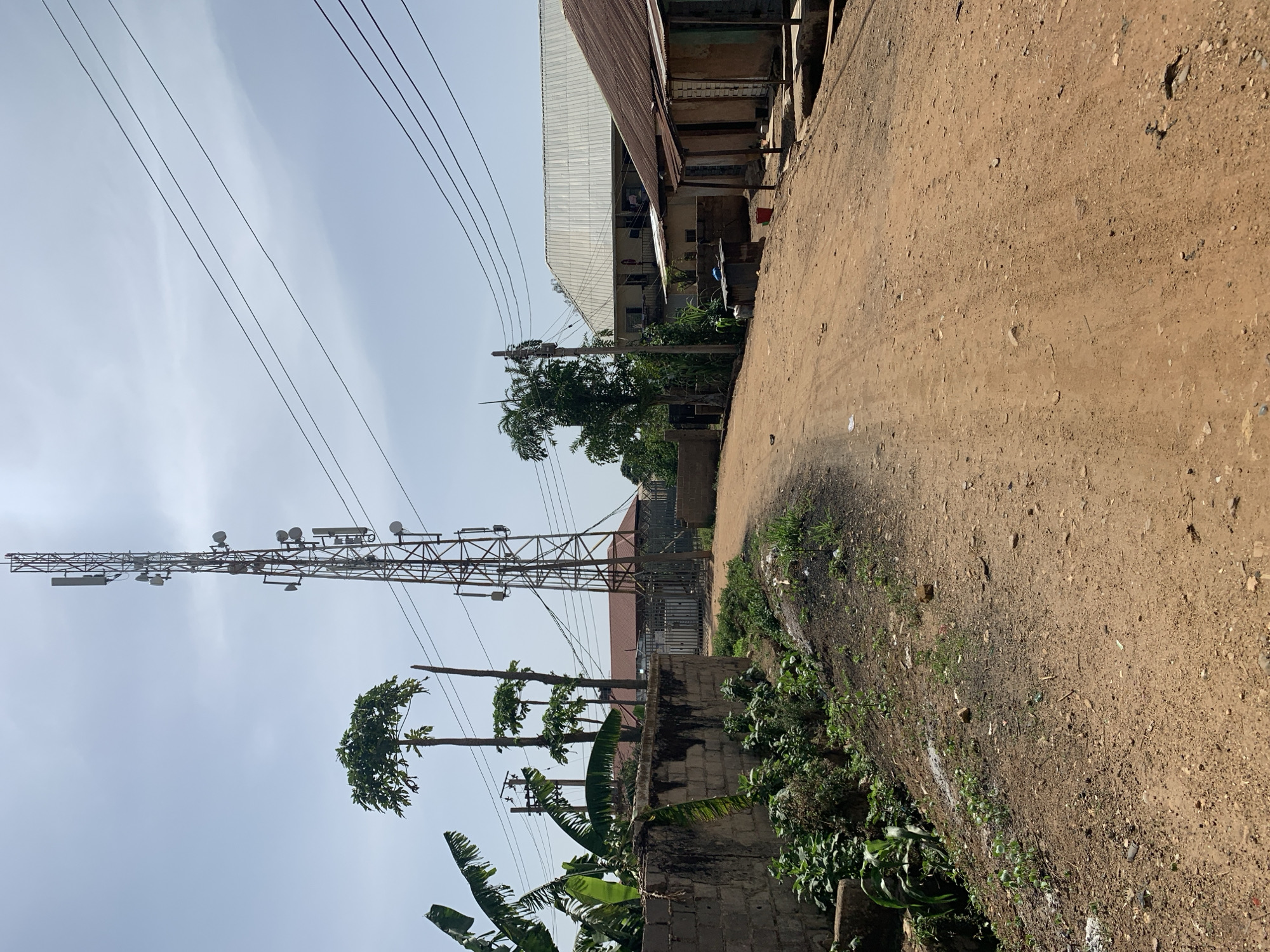 The image depicts a street scene in Rafin Par, Nigeria. A dirt road extends into the scene, bordered on the right by overgrown vegetation and a low brick wall. On the left, a tall telecommunications tower stands prominently, with power lines stretching across the sky. In the background, a building with a corrugated metal roof is visible. The sky is overcast. No people are visible in the image.