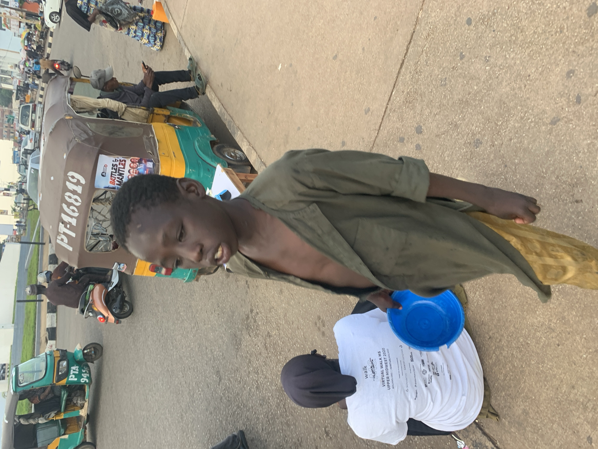 In Barakin Lerawi, Nigeria, a boy in a green shirt and gold pants stands near a blue bowl. He is the foreground subject, with a woman in a white shirt with "WALK MS UPPER MIDWEST 2020" printed on it sitting behind him. A tuk-tuk vehicle is parked nearby. The background shows a street with other vehicles and pedestrians. The scene appears to be a public area in a Nigerian city.