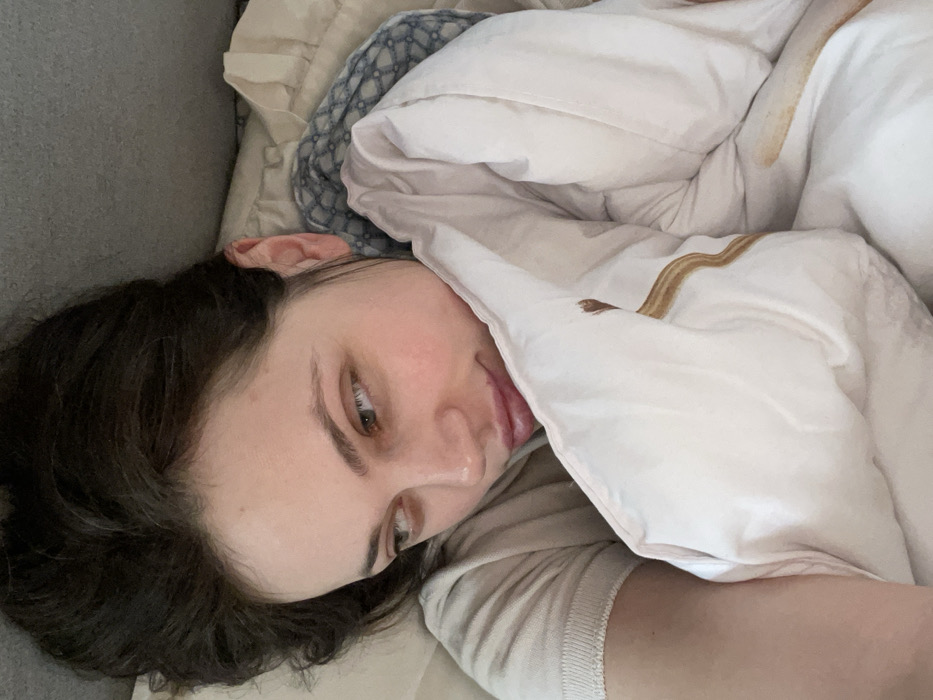 A close-up, high-angle shot depicts a female individual lying down indoors on her right side, her head oriented towards the upper left of the frame and her gaze directed to the left (viewer's right). She exhibits fair skin, dark brown hair, and light-colored eyes, with a neutral to slightly contemplative expression. She wears a light-colored, short-sleeved garment. Her head rests on a plain light-colored pillow, partially obscuring a blue and white patterned fabric behind it. A white duvet or blanket, marked with subtle light brown, wavy, abstract patterns or stains, covers a portion of her body. The immediate background is a plain light grey surface. This scene is situated in Monsummano Terme, Italy.