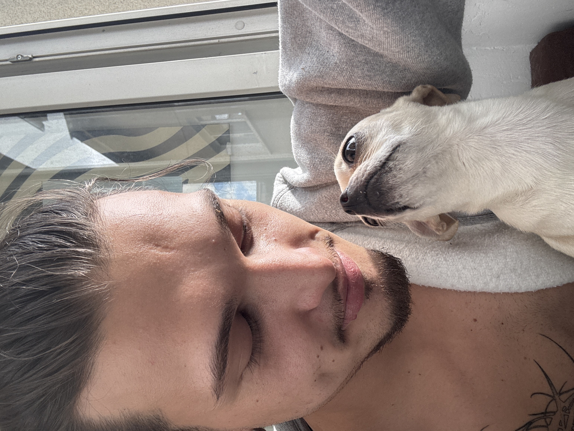 A close-up, waist-up, selfie-style shot captures a young person and a small dog on what appears to be a balcony. The person, with dark hair pulled back and a light beard, is looking down at the dog, their eyes closed. The dog, a light-colored chihuahua, is looking up at the person, its mouth slightly open. Both are wearing casual clothing. The background shows a window with blinds and a wall. The scene is illuminated by natural light, suggesting daytime. The location of Sofia, Bulgaria, is not discernible from the image itself.