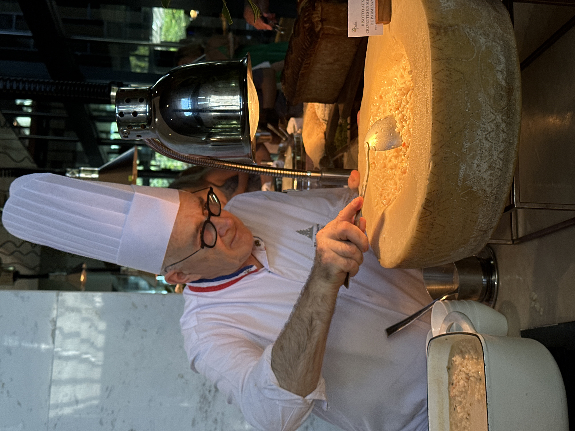 In Nusa Dua, Indonesia, a chef in a tall white hat and glasses, wearing a white uniform with red, white, and blue stripes on the collar, stands beside a large wheel of cheese. He is using a tool to scoop out a portion of the cheese under the light of a metal lamp. Part of a menu card is visible on top of the cheese wheel. A serving container is placed beneath the cheese.