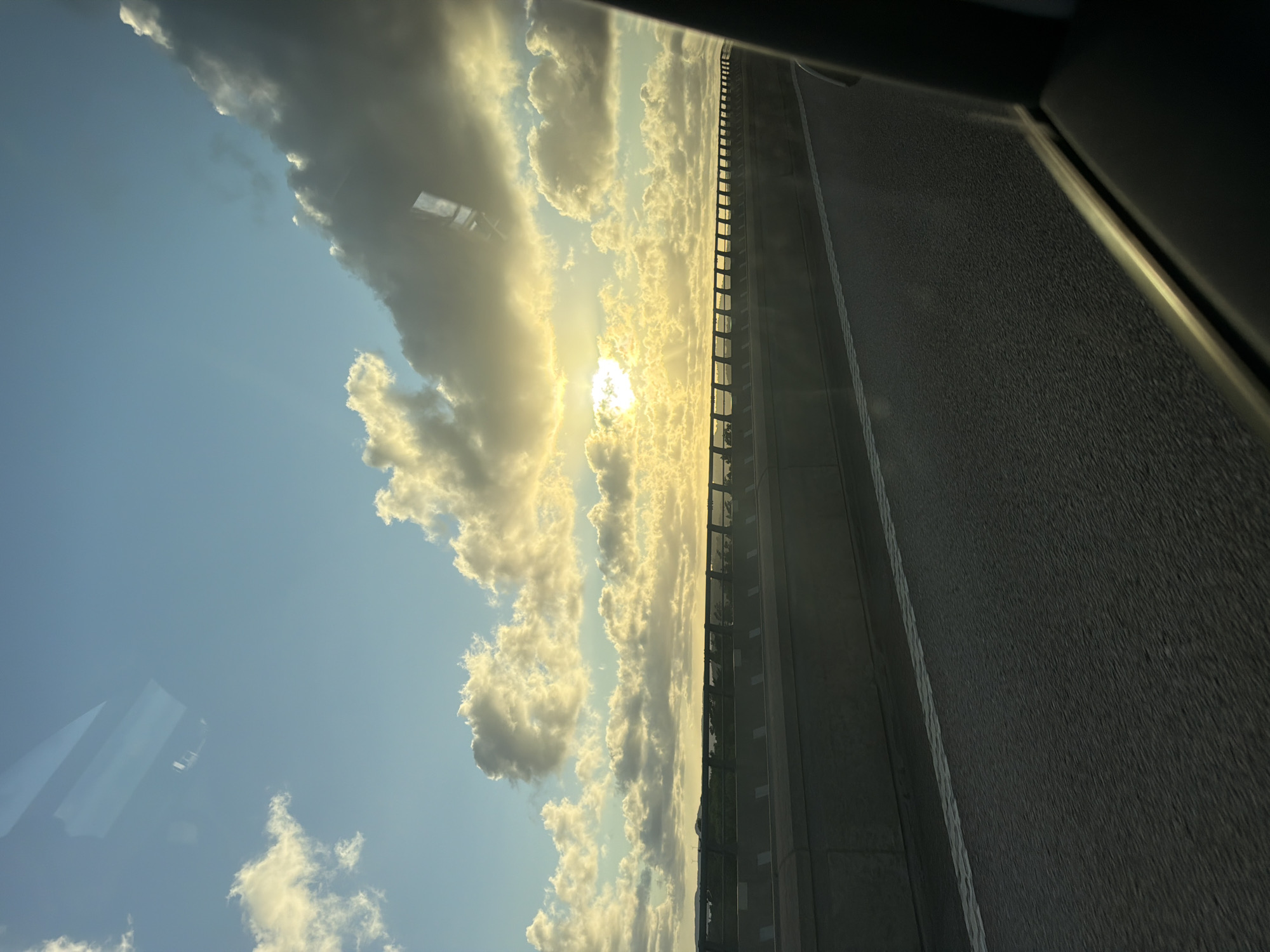 The image depicts a roadside view with a sunset in the background. The foreground shows a grey asphalt road with a white line marking the lane. To the left, a concrete barrier with a black metal railing runs along the edge of the road. Beyond the barrier, a distant landscape with trees and some structures is visible. The sky is filled with a mixture of blue sky and fluffy, golden-lit clouds, with the sun setting behind a large cloud formation. Reflections from a window are visible in the upper part of the image, obscuring some of the sky. The scene appears to be a highway or an elevated road, possibly in or near Ödsmål, Sweden, given the presence of wind turbines in the distance. There are no people visible in the image.