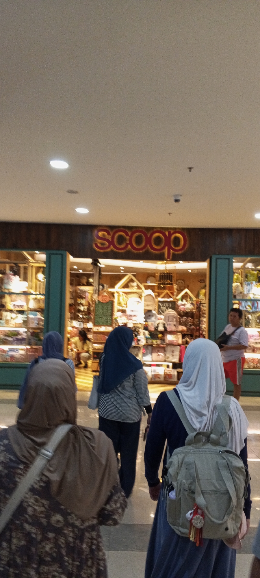 In a shopping mall in Depok, Indonesia, a group of individuals are seen walking towards a store labeled "scoop." The store's facade is adorned with warm, illuminated lettering, and through its display windows, a variety of items are visible, suggesting it is a retail establishment. The floor is a polished, light-colored material, and overhead lighting fixtures are integrated into the ceiling. Among the individuals, three women are prominently featured in the foreground, all wearing hijabs and modest clothing. One woman in the rear wears a brown patterned dress and a beige hijab. To her left, another woman is clad in a blue hijab and a striped long-sleeved shirt. A third woman, further to the right, wears a light blue hijab, a dark blue long-sleeved top, and a light-colored backpack adorned with decorative tassels. In the background, a man in a light purple shirt and red shorts stands near the entrance of the "scoop" store. The overall scene depicts a casual outing in a brightly lit indoor commercial space.