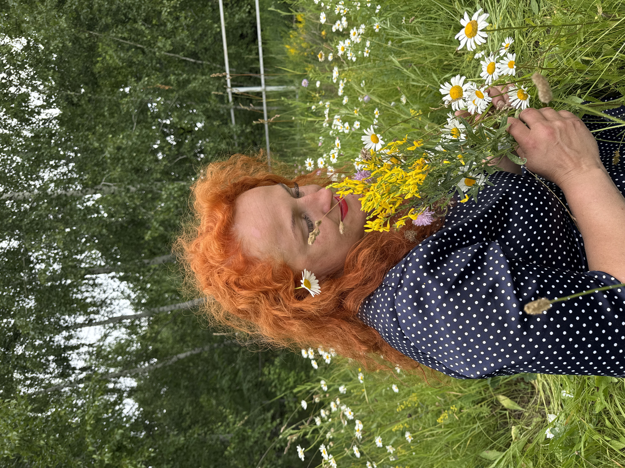 A woman with bright red, curly hair is lying down in a grassy field with wildflowers. She is wearing a dark blue dress with white polka dots. Her eyes are closed, and she appears to be relaxed, with a slight smile on her face. She is holding a bouquet of yellow wildflowers and daisies. A single daisy is tucked into her hair above her ear.
The scene is outdoors, with a background of green trees. The lighting suggests it is daytime, likely a sunny day given the vibrant colors of the flowers. The overall mood is peaceful and serene. There are no visible text elements in the image. The location context is Privolzhsky, Russia.