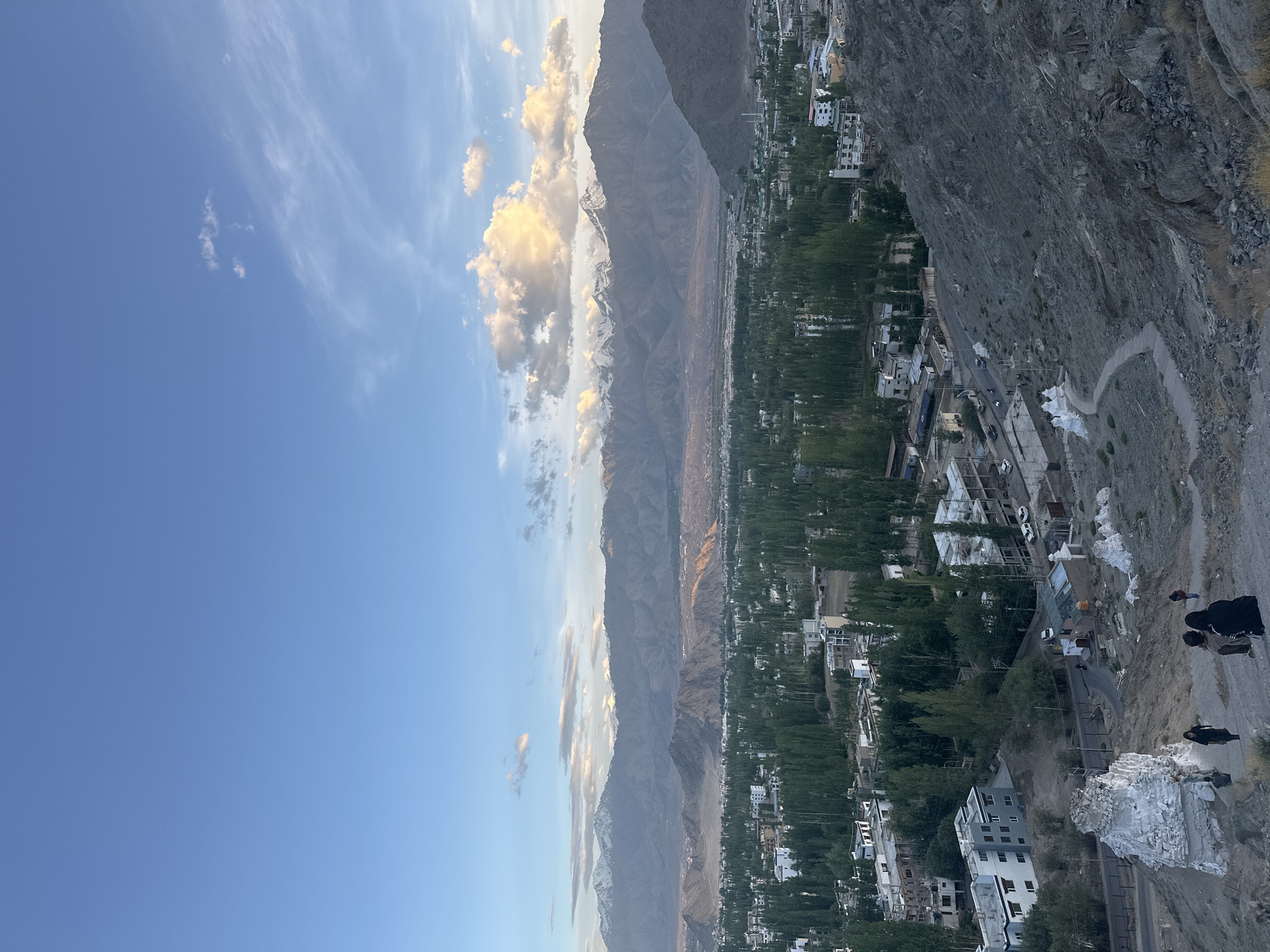 The image depicts an elevated, wide-angle view of the city of Leh, India, likely taken from a hilltop vantage point. The foreground features a steep, rocky slope with a few scattered white stupas and a pathway winding down towards the city. Several individuals, dressed in casual clothing, are visible walking on the slope, some of them appearing to be tourists.
In the midground, the city of Leh unfolds, characterized by a dense collection of buildings interspersed with numerous tall, slender trees. The architecture appears to be a mix of modern structures with white facades and balconies, and older, more traditional buildings. Roads with a few vehicles, including cars and what appears to be a motorcycle, are visible within the city.
The background is dominated by a dramatic mountain range, with snow-capped peaks piercing the sky. The lower slopes of these mountains are a muted brown and grey, while the higher elevations are covered in snow, reflecting the soft light of what appears to be late afternoon or early evening. The sky above is a gradient of blue, with scattered white and golden clouds, some of which are illuminated by the setting sun. The overall impression is one of a vast, mountainous landscape encompassing a bustling urban center.