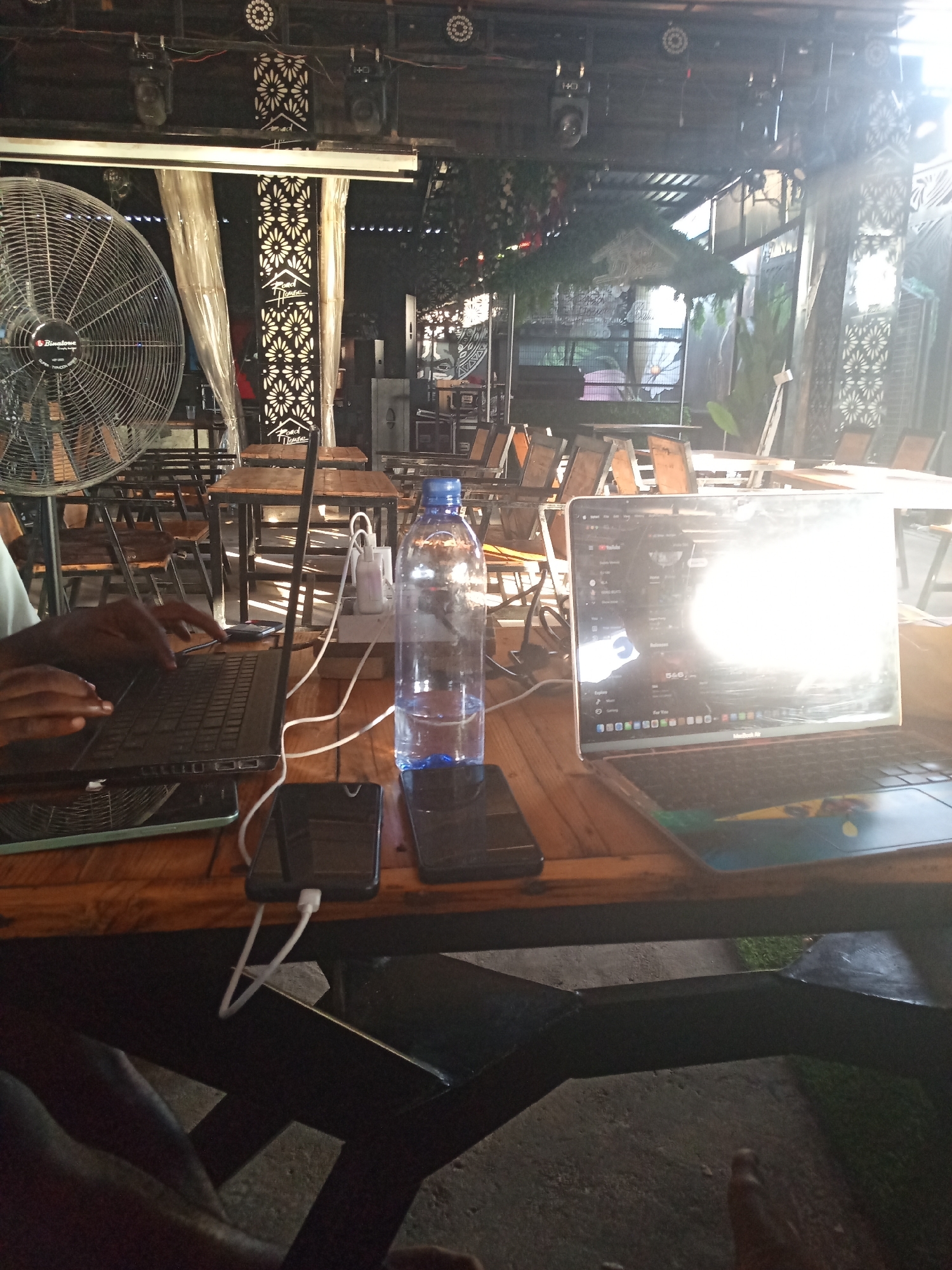 This image captures an indoor or semi-outdoor commercial establishment, identified by "Road House" branding on decorative panels, in Howayi, Nigeria. The scene is illuminated by strong natural light from the right and supplementary artificial lighting from fixtures mounted on an overhead dark metal beam structure.
In the foreground, a dark wooden table occupies the lower half of the frame. On this table, from left to right, are two laptops, a water bottle, and multiple smartphones. A person's dark-skinned hands are visible on the left, actively typing on a black laptop. A tablet with a greenish-blue case is partially visible beneath this laptop. To the right of the first laptop, a clear plastic water bottle with a blue cap, approximately half-full, stands upright. Further right, a silver/grey MacBook Air laptop is open, its screen displaying what appears to be a YouTube interface, though the screen is largely obscured by severe sun glare. The MacBook's keyboard features a colorful sticker. Between the laptops and to the right of the left laptop, a white power strip or multi-port charger is connected to several white charging cables. At least two black smartphones are on the table, one explicitly connected to a white charging cable. A person's leg and foot are partially visible in the bottom right corner of the frame.
The background reveals rows of dark wooden tables with metal frames and accompanying chairs, arranged in a seating area. A large, metallic, round stand fan branded "BINGSTONE" is positioned on the far left. The overhead structure includes exposed metal beams, electrical wiring, spotlights, and decorative LED lights. Two prominent decorative panels featuring a white floral pattern against a dark background, each bearing the text "Road House," are visible centrally in the mid-ground. Additional decorative elements include draped translucent fabric and some artificial or real greenery. The floor visible beneath the table structure appears to be concrete, with patches of green vegetation further in the background.