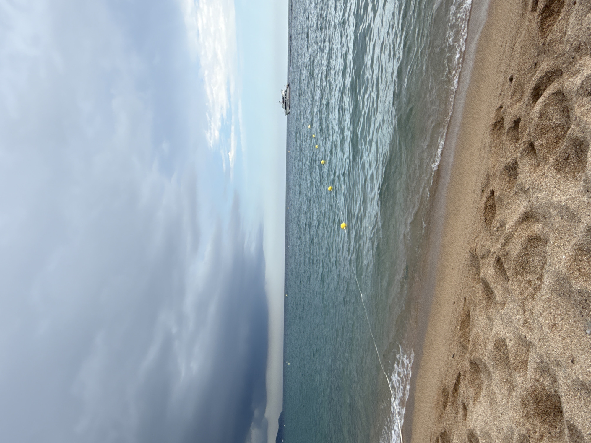 The image depicts a beach scene in Blanes, Spain, under a cloudy sky. The foreground features a sandy beach with visible footprints. The sea occupies the middle ground, with gentle waves lapping at the shore. A white rope stretches from the beach into the water, marked by yellow buoys. In the distance, a white boat with dark lettering is visible on the horizon, moving from left to right. The sky is filled with dark, heavy clouds on the left, transitioning to lighter, scattered clouds on the right. A faint landmass is visible on the far left horizon.