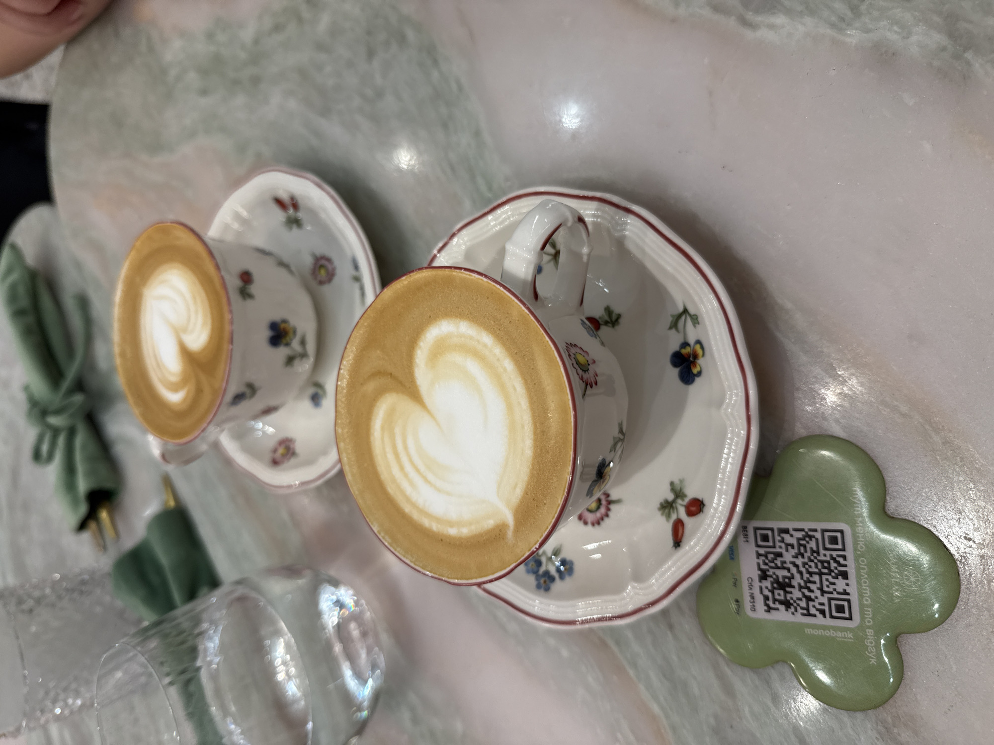 Two cups of cappuccino with latte art in the shape of a heart are placed on saucers. The cups and saucers are white with a floral pattern and a red rim. One cup is in the foreground, and the other is in the background, slightly to the right. A glass of water is to the left of the foreground cup. A green clover-shaped item with a QR code is on the table to the left of the foreground cup. The table appears to be made of marble with green and white veining. The setting is likely a cafe or restaurant in Kyiv, Ukraine, as indicated by the QR code which contains Ukrainian text and references "monobank".