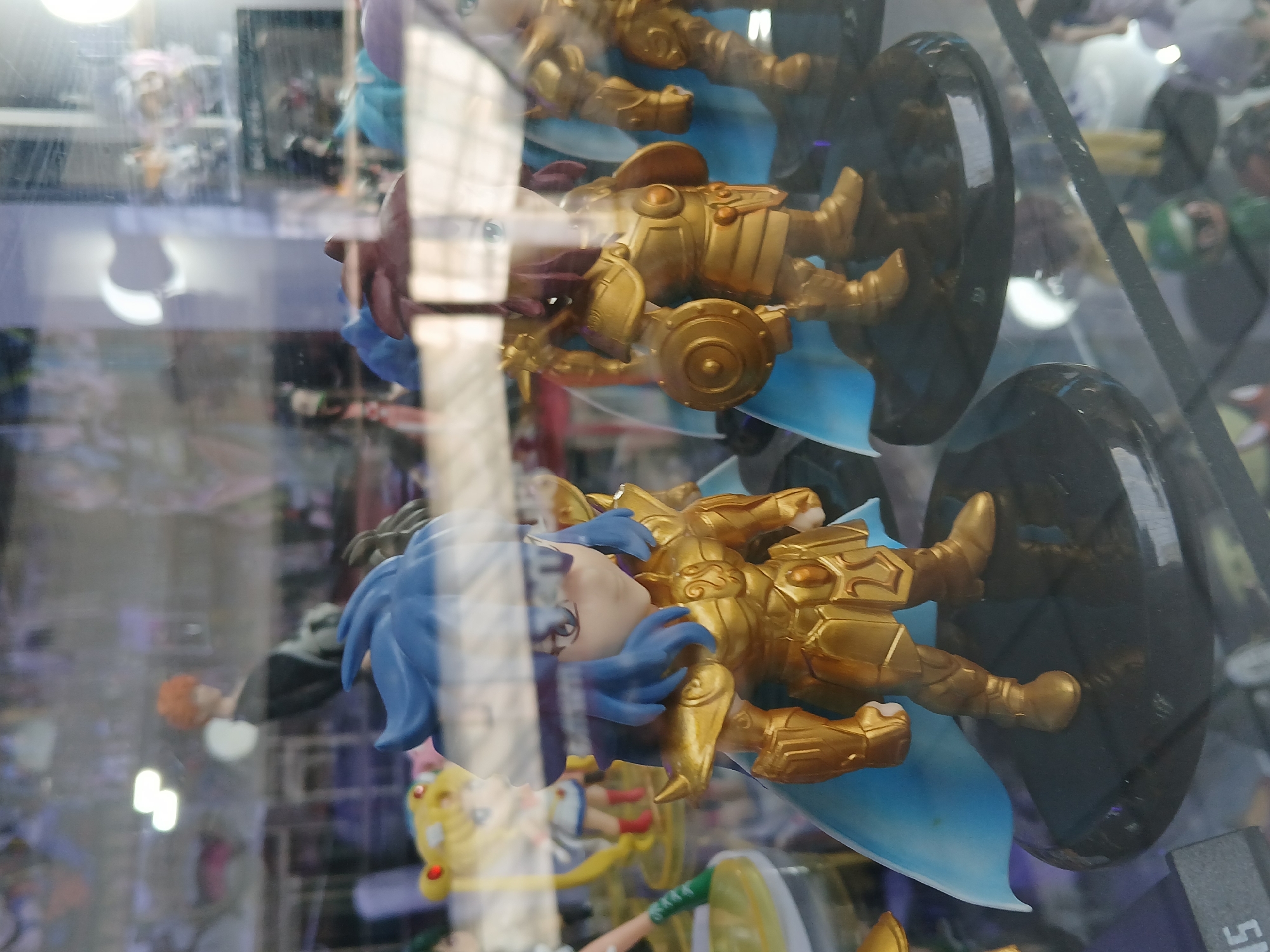In a display case, presumably in a retail setting in Ciudad Guayana, Venezuela, three anime-style figurines are arranged vertically. The figurines appear to be gold-colored characters in armor with flowing blue capes. The bottom figurine has blue hair, and the middle figurine has brown hair. The figurines are mounted on dark-colored bases. The display case is transparent, and reflections of the surrounding environment can be seen.