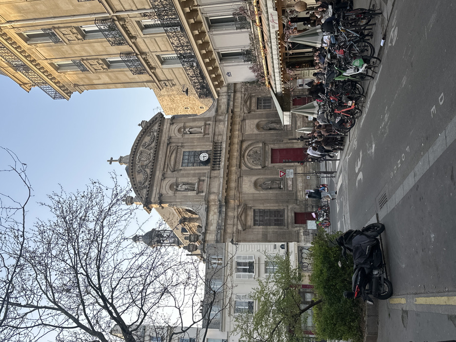 The image depicts the Église Saint-Paul-Saint-Louis, a Baroque church located at 99 Rue Saint-Antoine, 75004 Paris, France, within the Marais district.
The church's light-colored stone facade dominates the center, featuring two main levels with statues in niches and a prominent clock on the upper level, capped by a cross at its apex. A dark, ornate bell tower with a cupola rises from the church's roofline on the left. The main entrance consists of large red wooden doors, fronted by a low metal fence and a "Cédez le Passage" (yield) sign.
To the immediate right of the church stands a multi-story, cream-colored Haussmannian building with numerous windows and balconies adorned with black iron railings. At its ground level, a café identified as "Café Jourdain" features an outdoor seating area under beige awnings and white umbrellas. Approximately twenty individuals are seated at tables, engaged in social interaction. A dense line of parked bicycles, including several shared rental bikes, occupies the curb in front of the café.
To the left of the church, a lighter-colored, multi-story building with white window shutters is partially visible, behind a small green planter and sparse, bare-branched trees in the foreground. A black scooter or motorcycle with a dark cover is parked on the street in the left foreground.
The foreground consists of an asphalt street with faded white markings, including a reversed "LONDRES" and an arrow, indicating traffic flow. A few pedestrians are visible crossing the street or walking on the sidewalk near the church entrance. The sky is clear and light blue, suggesting fair weather.