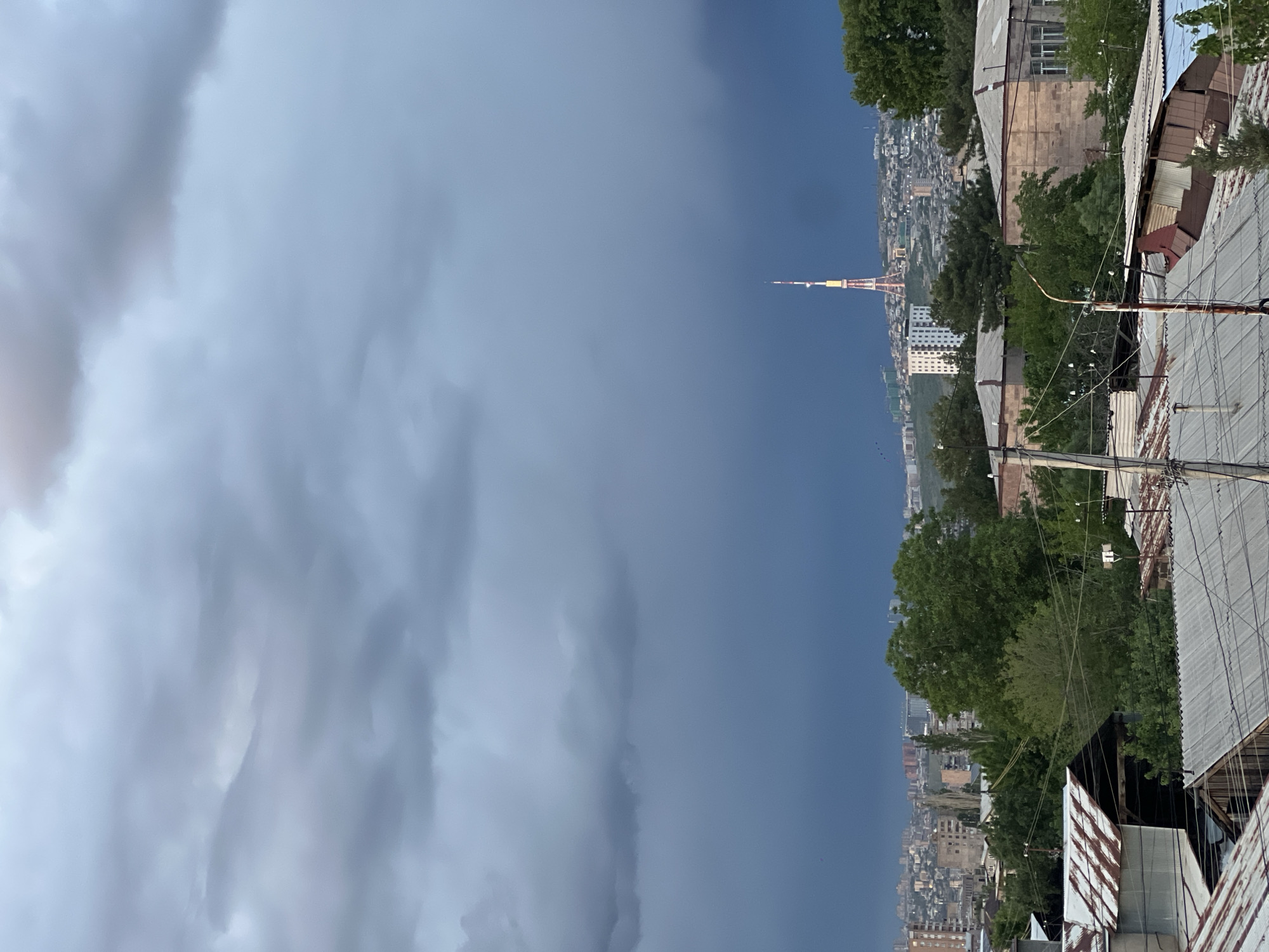 A cityscape view from Yerevan, Armenia, features a tall television tower reaching into a stormy sky. The dense clouds suggest a coming or recent rain. Buildings of varying heights and designs cascade down the hillside to the horizon, punctuated by green trees. In the foreground, the corrugated iron roofs of older buildings with power lines running alongside and above are visible. The light levels suggest it is late afternoon or early evening.