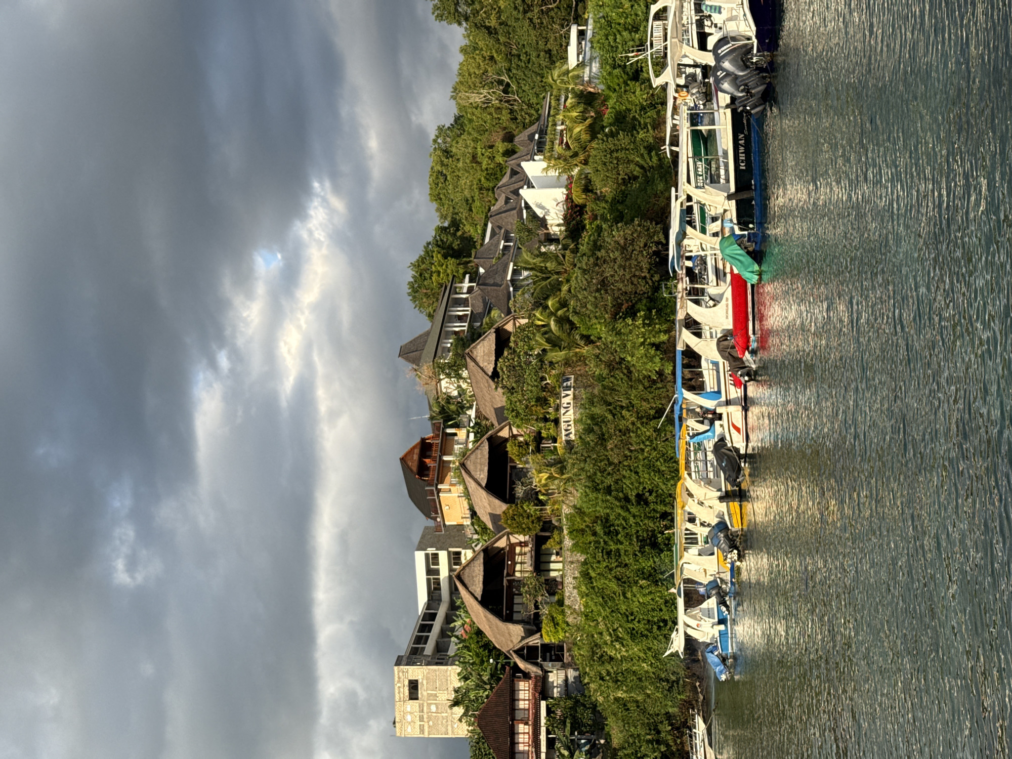 The image captures a view of Nusa Penida, Indonesia, featuring buildings and a row of boats along the waterfront. Several structures with traditional thatched roofs are visible amidst lush greenery on the hillside. Other buildings with more modern architectural designs are also present. Adjacent to the green vegetation, a series of boats are docked, varying in size and color, each equipped with outboard motors. The water surface is textured with ripples. Overhead, the sky is overcast with dramatic cloud formations, casting shadows across the scene.