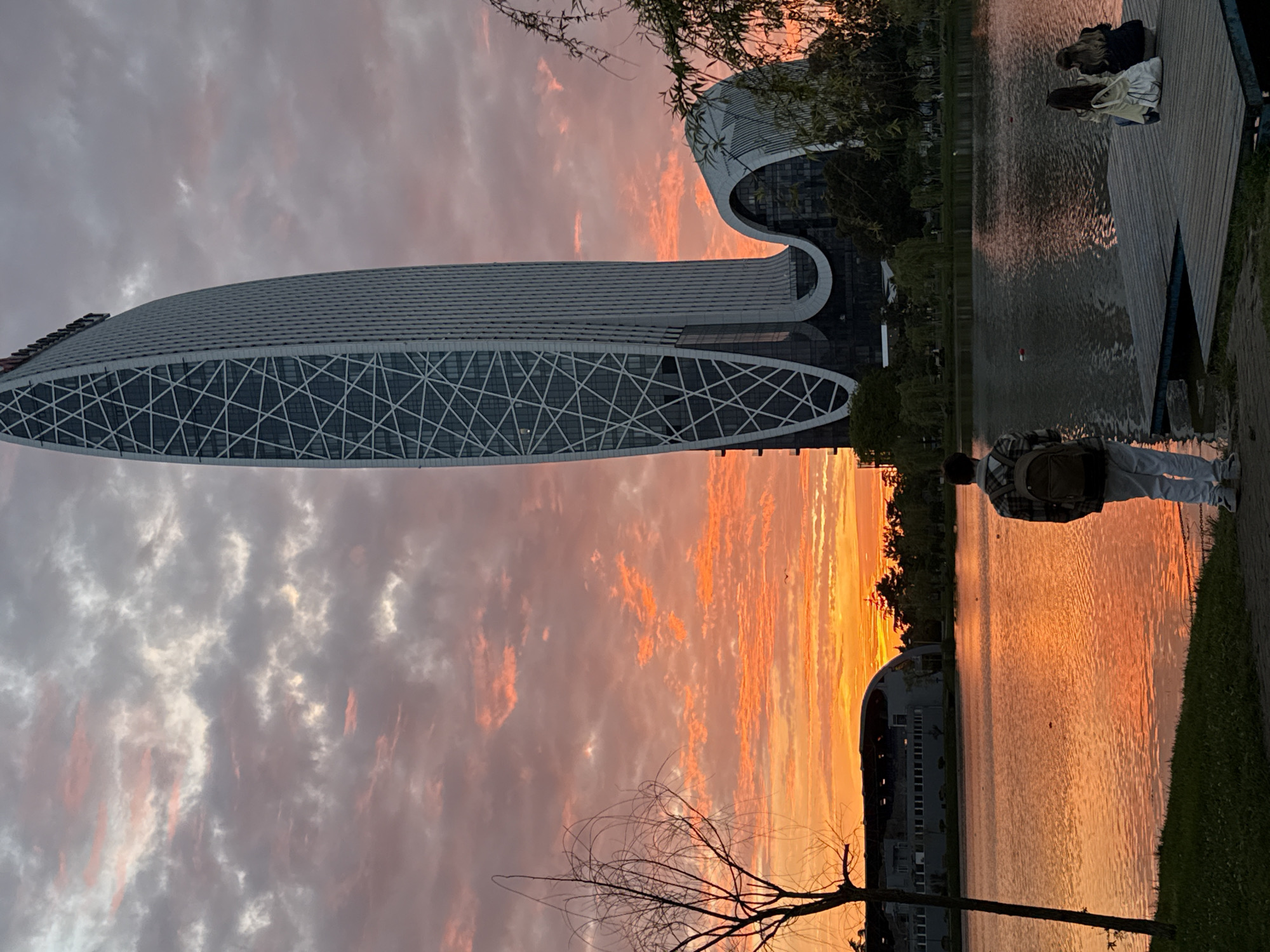 Captured at sunset in Batumi, Georgia, the image features a modern architectural building with a sleek, elongated design, seemingly reflected in the waters of a nearby lake. The sky is ablaze with fiery hues of orange and pink, casting a warm glow over the scene. Several people are present, including two individuals sitting on a wooden dock by the lake and another person walking along the edge of the lake near the buildings. There's a tree with bare branches silhouetted against the vibrant sunset, adding a natural element to the urban setting.