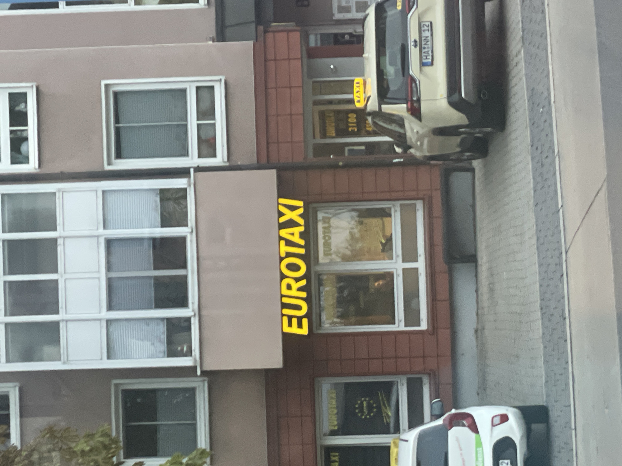 The image shows the exterior of a building in Hagen, Germany, with a prominent yellow "EUROTAXI" sign illuminated above a storefront. Two taxis are visible in front of the building. One is a white car parked on the left, and the other is a cream-colored SUV parked on the right, featuring a yellow taxi sign on its roof and a German license plate that reads "HA NN 12". The building has several windows, some with blinds drawn, and a brick facade on the lower level. The overall impression is a street-level view of a taxi service establishment.