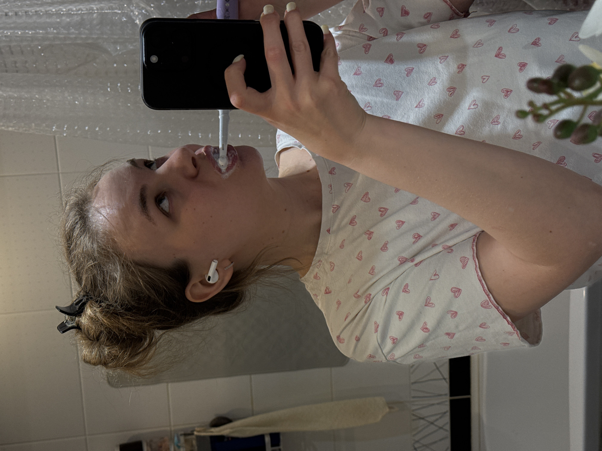 A woman is brushing her teeth in what appears to be a bathroom. She's holding a phone in front of her, likely taking a selfie or recording a video. She is wearing pajamas with a heart pattern. There's a shower curtain visible in the background, along with bathroom tiles and a cabinet. There are flowers and other objects also in the background of the photo. There is no information to infer the location in the city of Vladimir, Russia.
