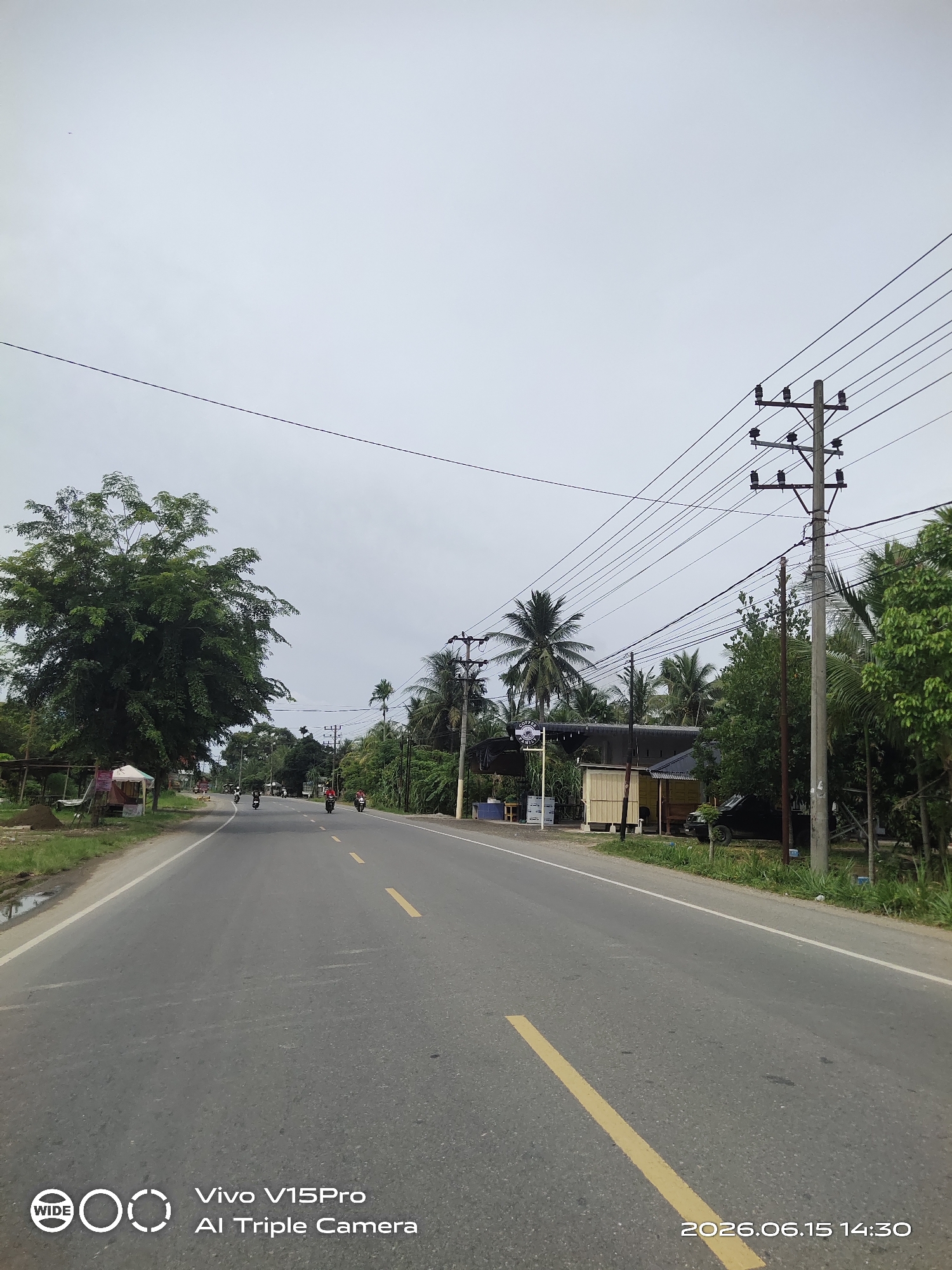 The image shows a straight, asphalt road with a yellow dashed line dividing the lanes. The road is lined with lush green vegetation and palm trees on both sides. Several utility poles with power lines run along the right side of the road. In the distance, a few motorcycles are visible on the road, suggesting traffic. On the left side of the road, there is a small roadside stall with a white tent. The sky is overcast. The timestamp in the bottom right corner indicates the photo was taken on June 15, 2026, at 14:30. The Vivo V15Pro AI Triple Camera watermark is present in the bottom left corner. The location is identified as Cotpuuk, Indonesia.
