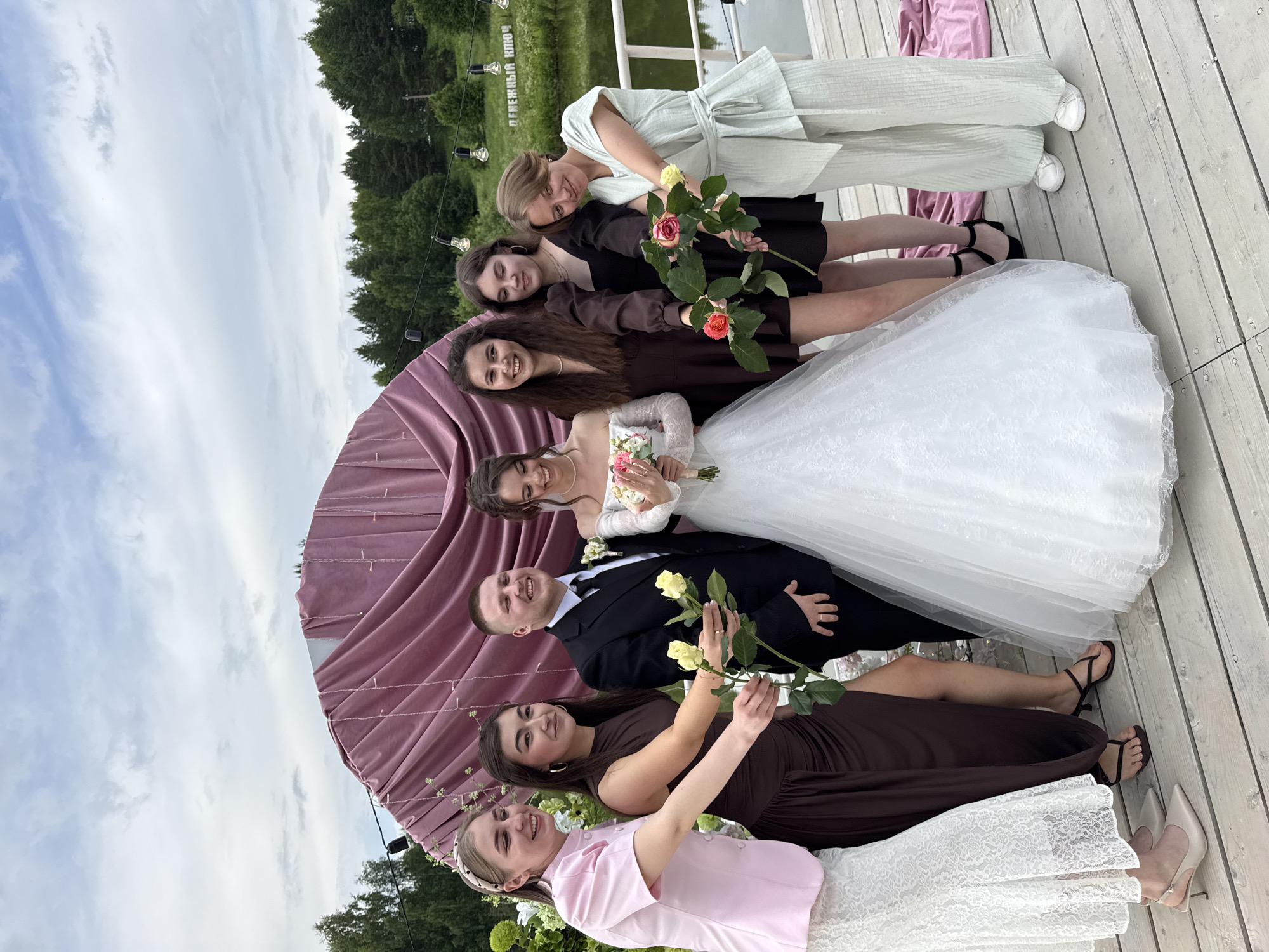 In Votkinsk, Russia, a wedding party is gathered on a wooden pier near a body of water. The bride, in a white lace wedding dress, stands between the groom, who is wearing a black suit, and several women. The bride is holding a bouquet of flowers, and the groom has a boutonniere. The women, dressed in various colored gowns and pantsuits, are also holding flowers. Behind them, a large, draped archway in a dusty rose color suggests a ceremony backdrop. String lights hang above the scene, and lush green trees and a cloudy sky form the background. The overall atmosphere is celebratory, with the wedding party smiling and posing for the photograph.