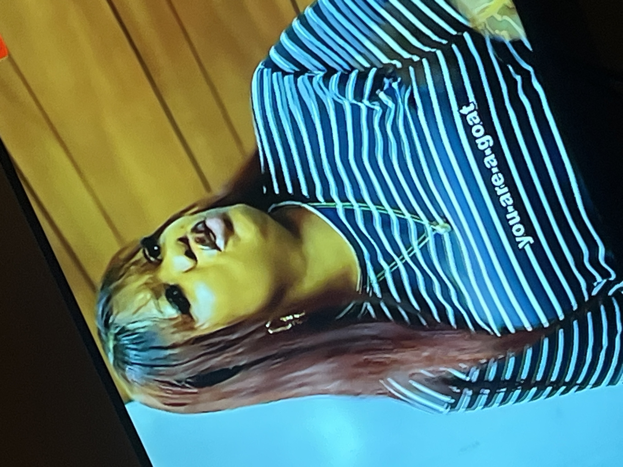 The image shows a woman with long, dark reddish-brown hair and bangs, wearing a horizontally striped t-shirt with blue and white stripes. The t-shirt has the text "you-are-a-goat" printed in white lettering. She is wearing hoop earrings and a necklace. Her facial expression appears to be one of speaking or making a statement. The background is a wooden-paneled wall with horizontal lines, and a bright blue area is visible on the left side of the image, possibly from a screen or lighting. The image quality is somewhat grainy, suggesting it may be a photograph of a screen. There is no information about the location in Gura Top, Nigeria.
