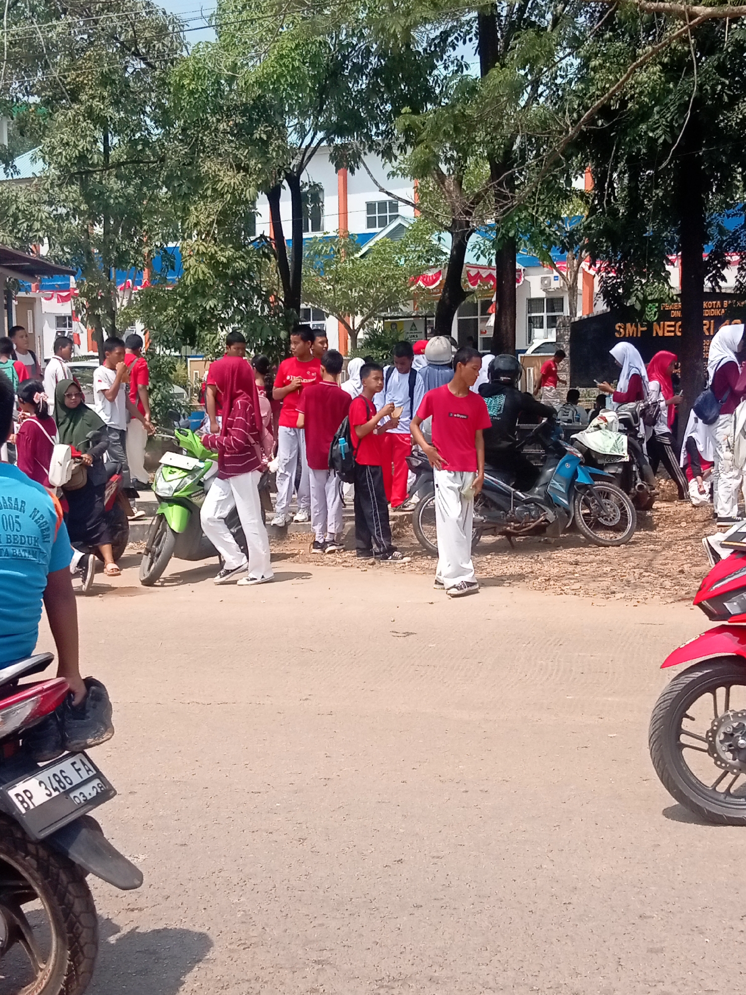 The image shows a group of students, some in uniform, gathered outside a building that appears to be a school. The building has a modern architectural style with white walls and blue accents. A sign on the building reads "SMP NEGERI" which translates to "Public Junior High School". The students are standing and interacting with each other, some holding mobile phones. Several motorcycles are parked in the foreground and middle ground, suggesting students may use them for transportation. The ground is paved with a dirt-like material, and trees line the background, providing shade. In the foreground, a blue-shirted person is partially visible from the back, sitting on a motorcycle. The overall scene depicts a typical day at a school in Batam, Indonesia, possibly during a break or after school hours. The license plate on the motorcycle in the foreground reads "BP 3486 FA", which indicates it is registered in the Riau Islands province, where Batam is located.